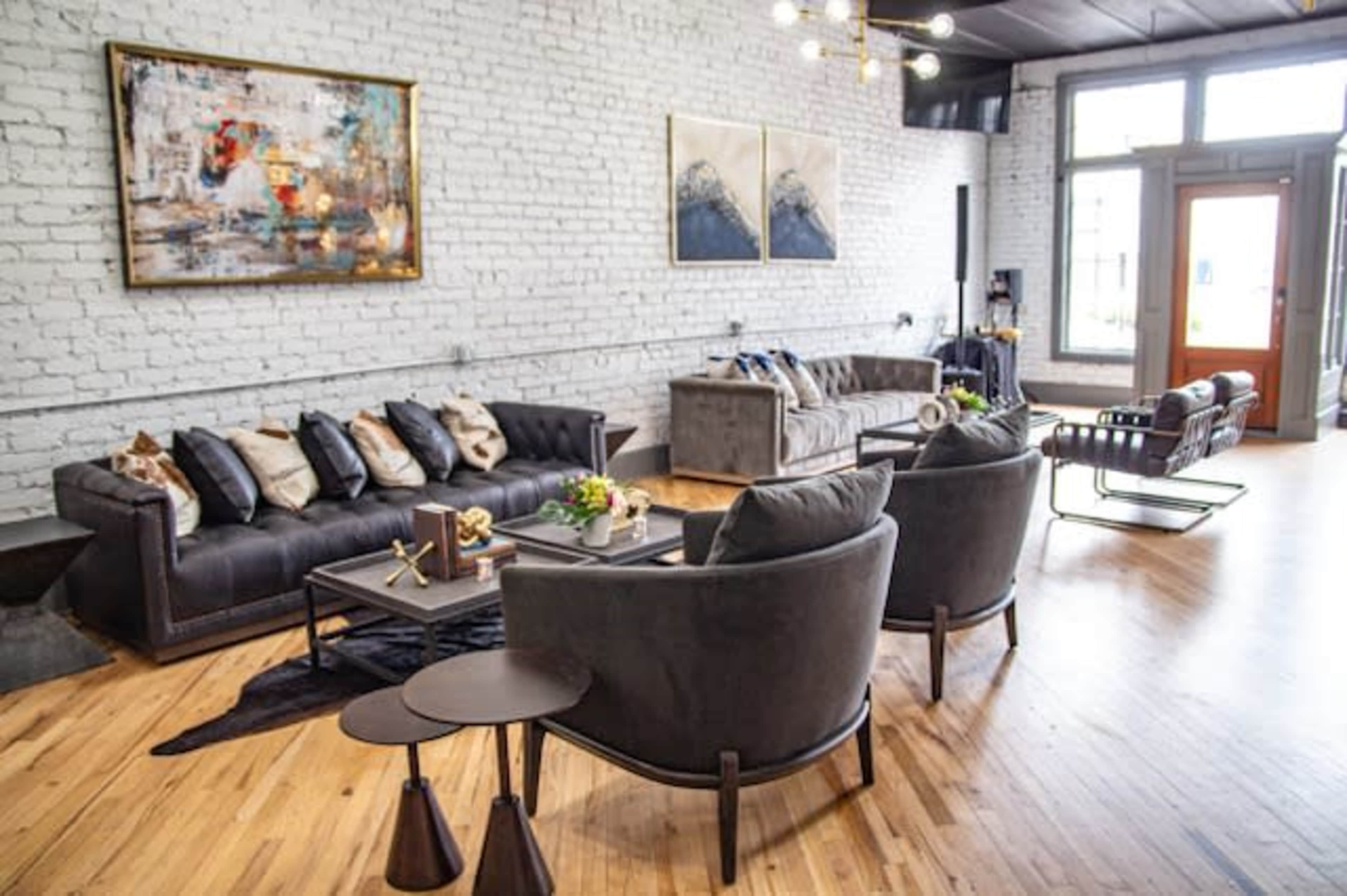 The image shows a modern living room featuring two sets of dark sofas, a coffee table with decorative items, and large windows with natural light streaming in.
