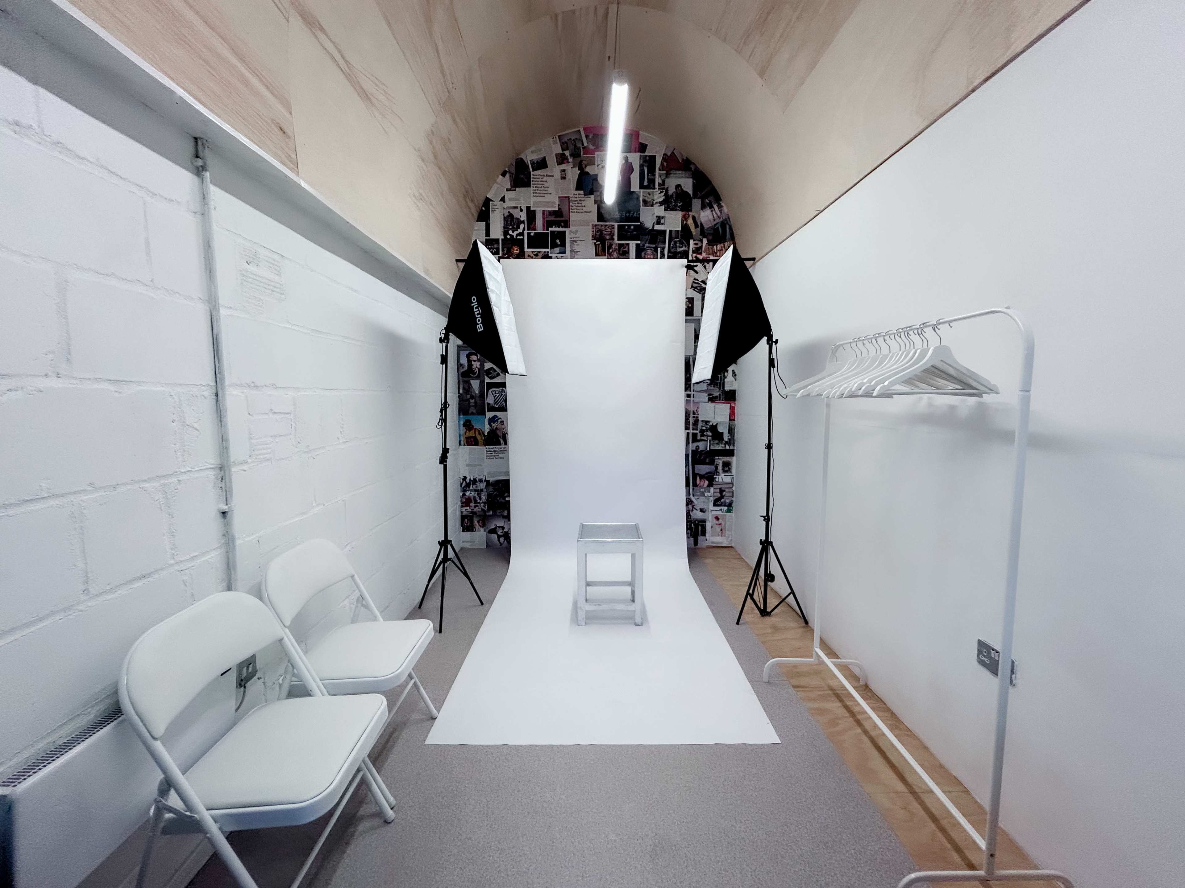 The image shows a minimalistic photography studio with a white backdrop, a stool in the center, two light stands, and a rack for clothing.