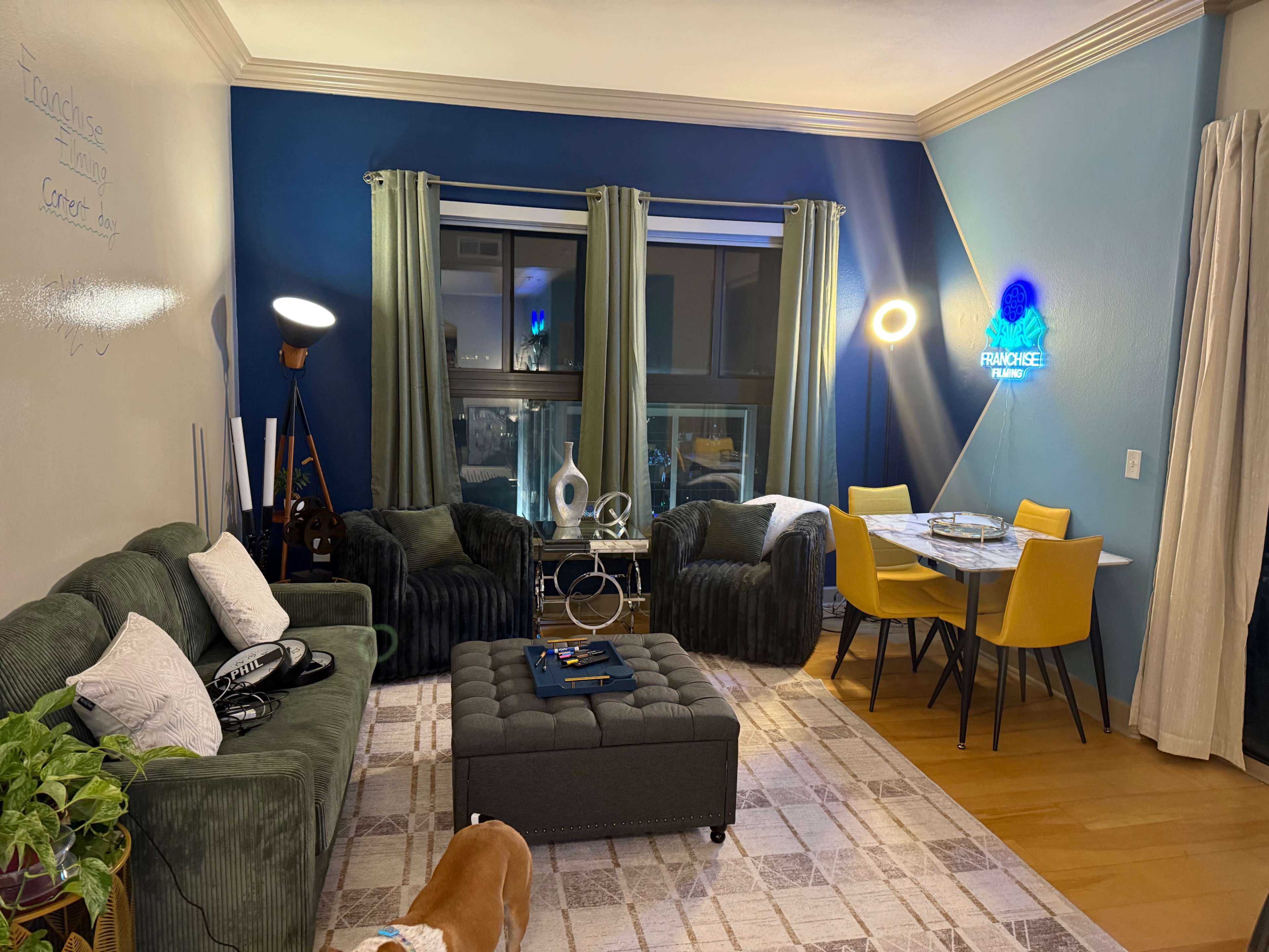 The image shows a modern living room with green and blue walls, two green armchairs, a gray ottoman, a dining table with yellow chairs, and large windows.