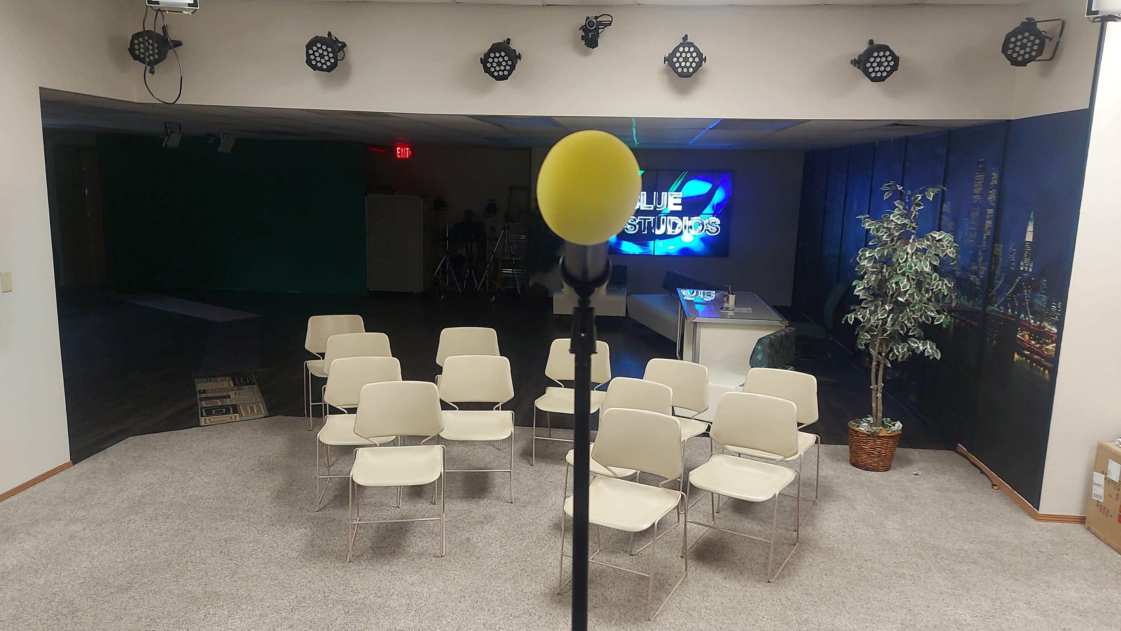 A microphone on a stand is positioned in front of empty chairs arranged in a studio setting with a backdrop displaying "BLUE STUDIOS."