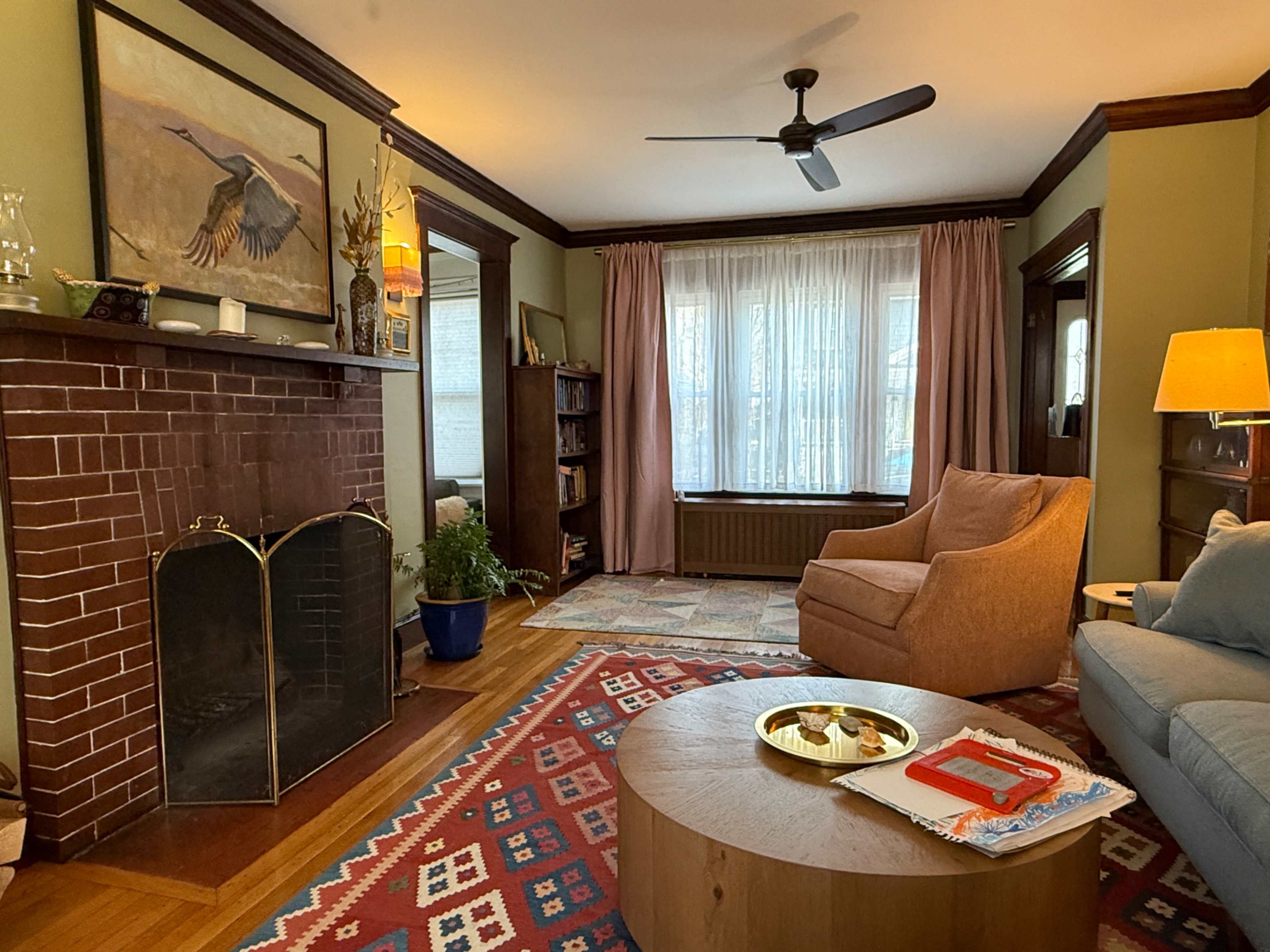 The image shows a cozy living room with a fireplace, a large window dressed with sheer curtains, and a combination of armchairs and a coffee table on a patterned rug.
