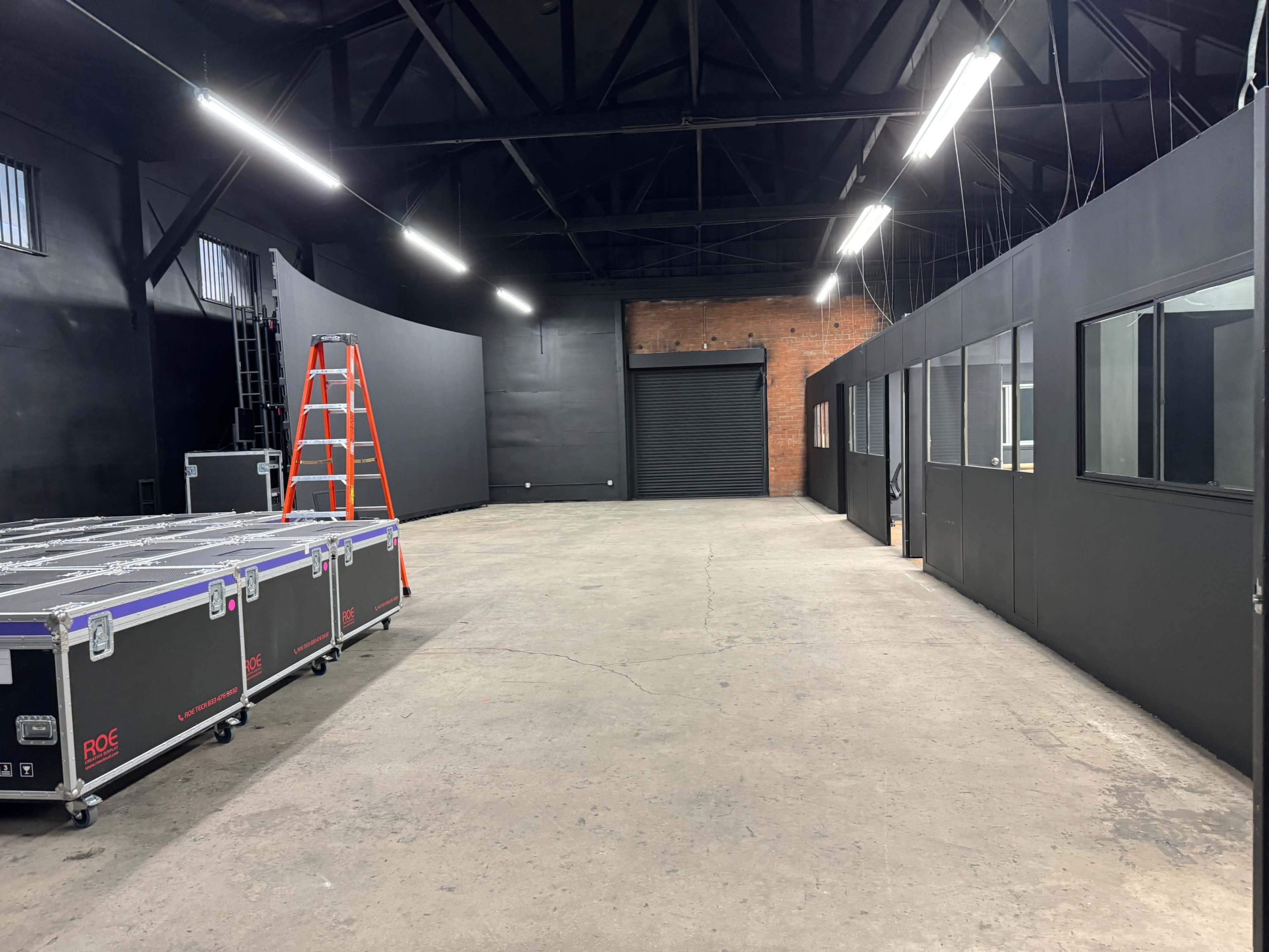 The image shows a spacious, industrial-style room with black walls, a concrete floor, a ladder against one wall, and equipment cases stacked on the floor.