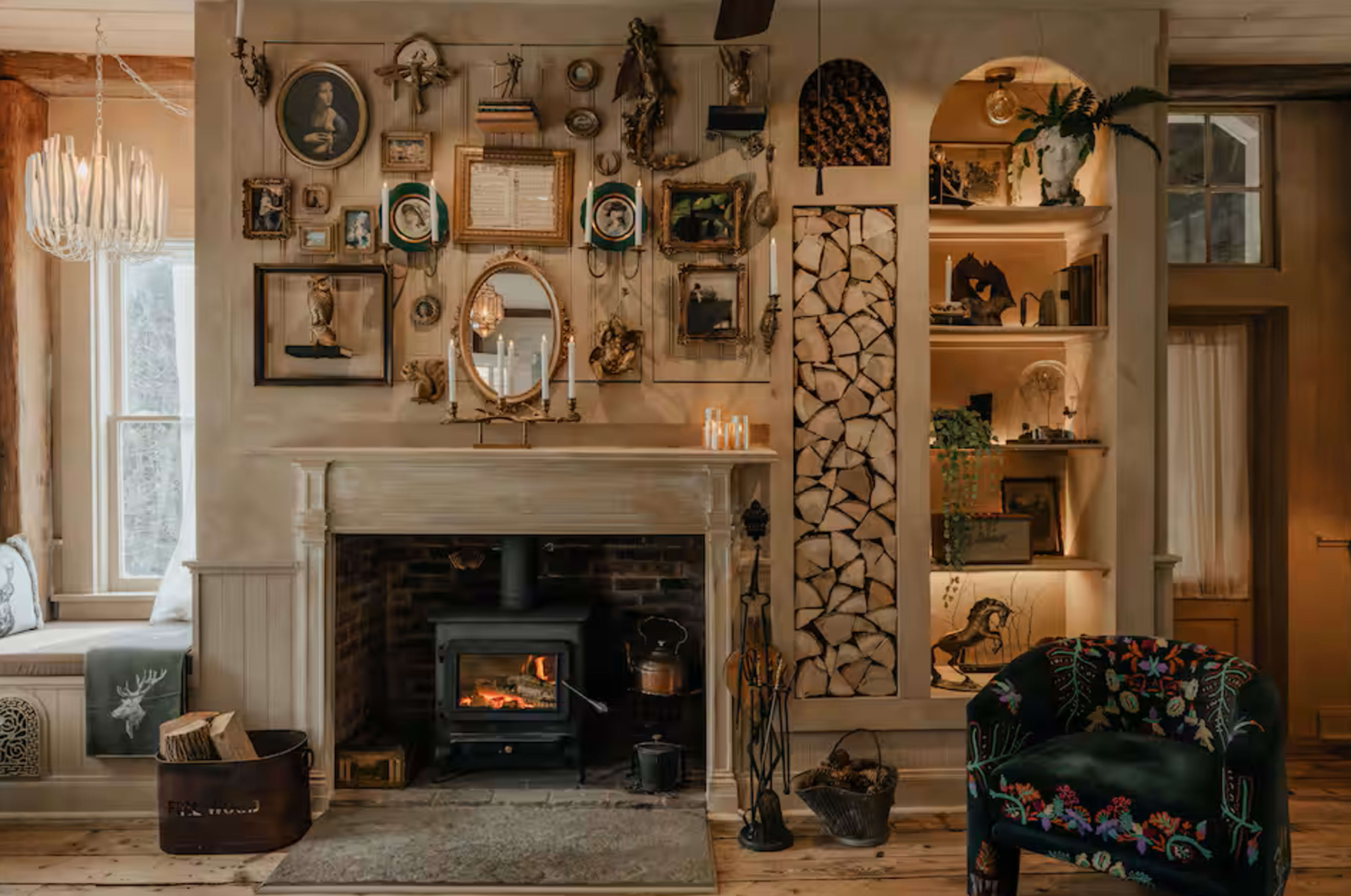 A cozy living room features a fireplace surrounded by an eclectic array of framed art and decorative items, along with a stacked wood display and a plush armchair.