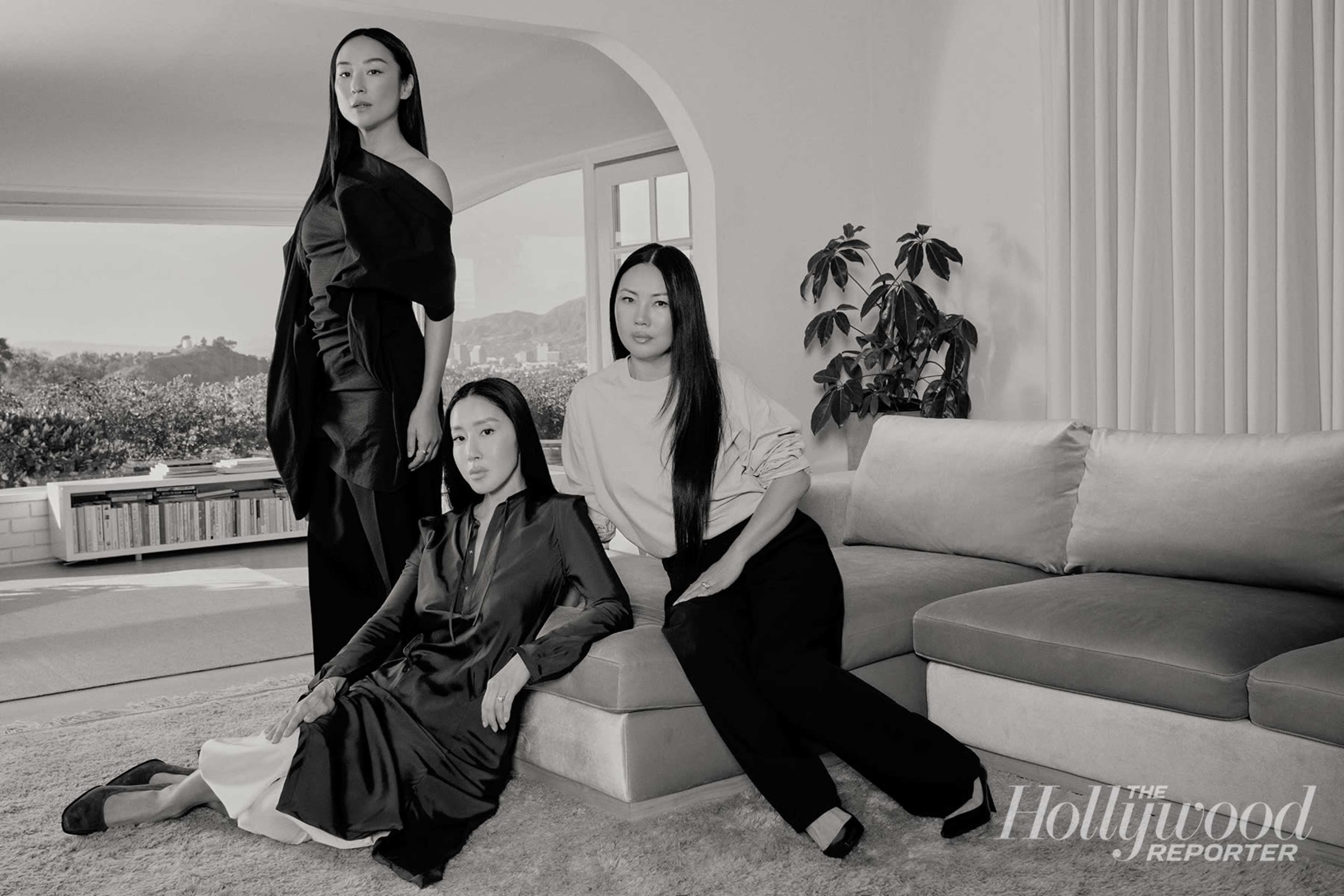 Three women pose in a modern living room, with one seated on a couch and the others standing behind her.