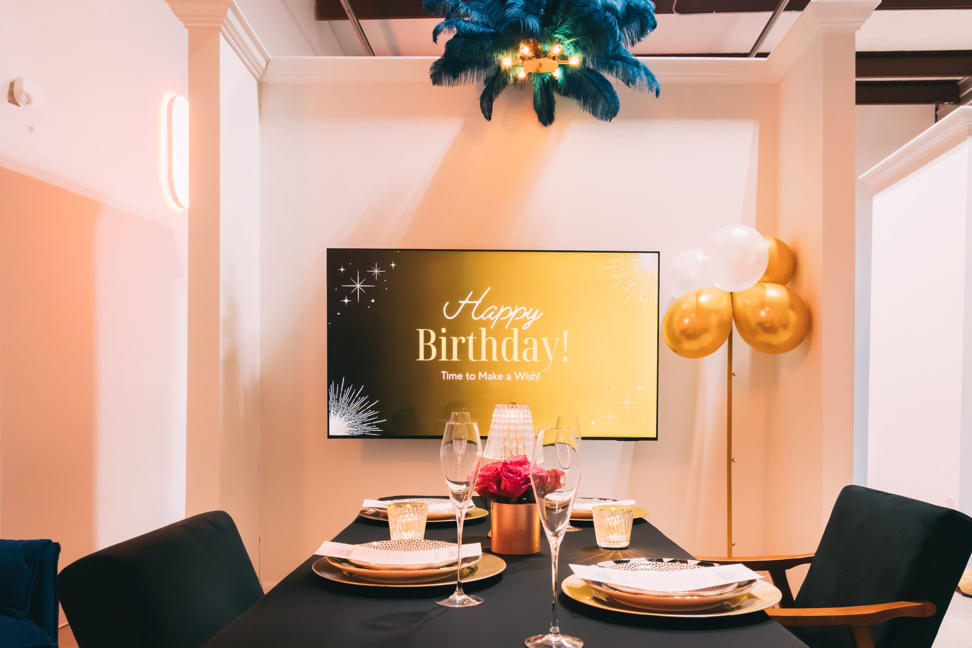A table is set for a birthday celebration in a brightly lit room, featuring a television displaying a festive birthday message along with gold and white balloons.