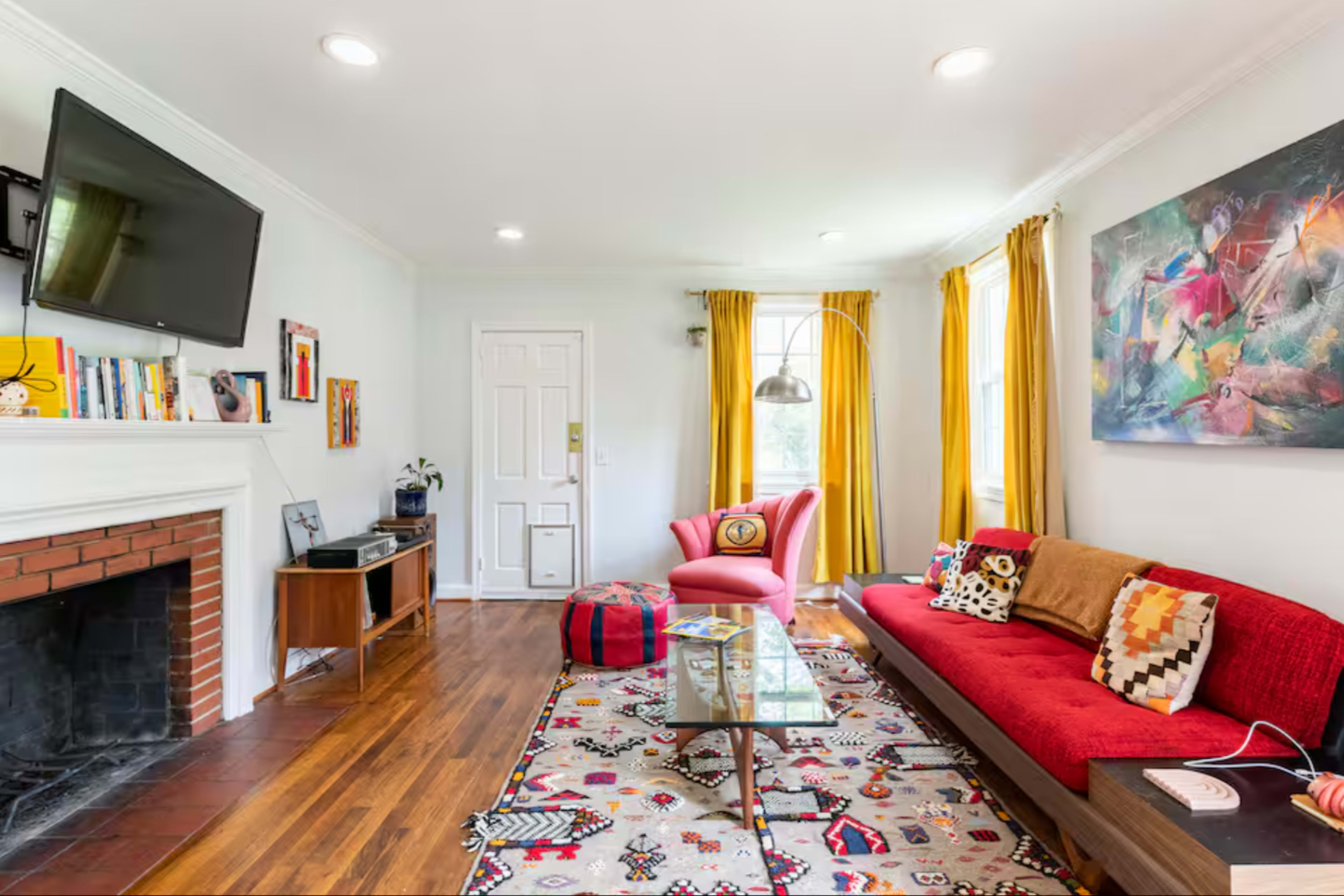 The living room features a red sofa, a pink chair, a coffee table, and a fireplace, with bright yellow curtains and a large television mounted on the wall.