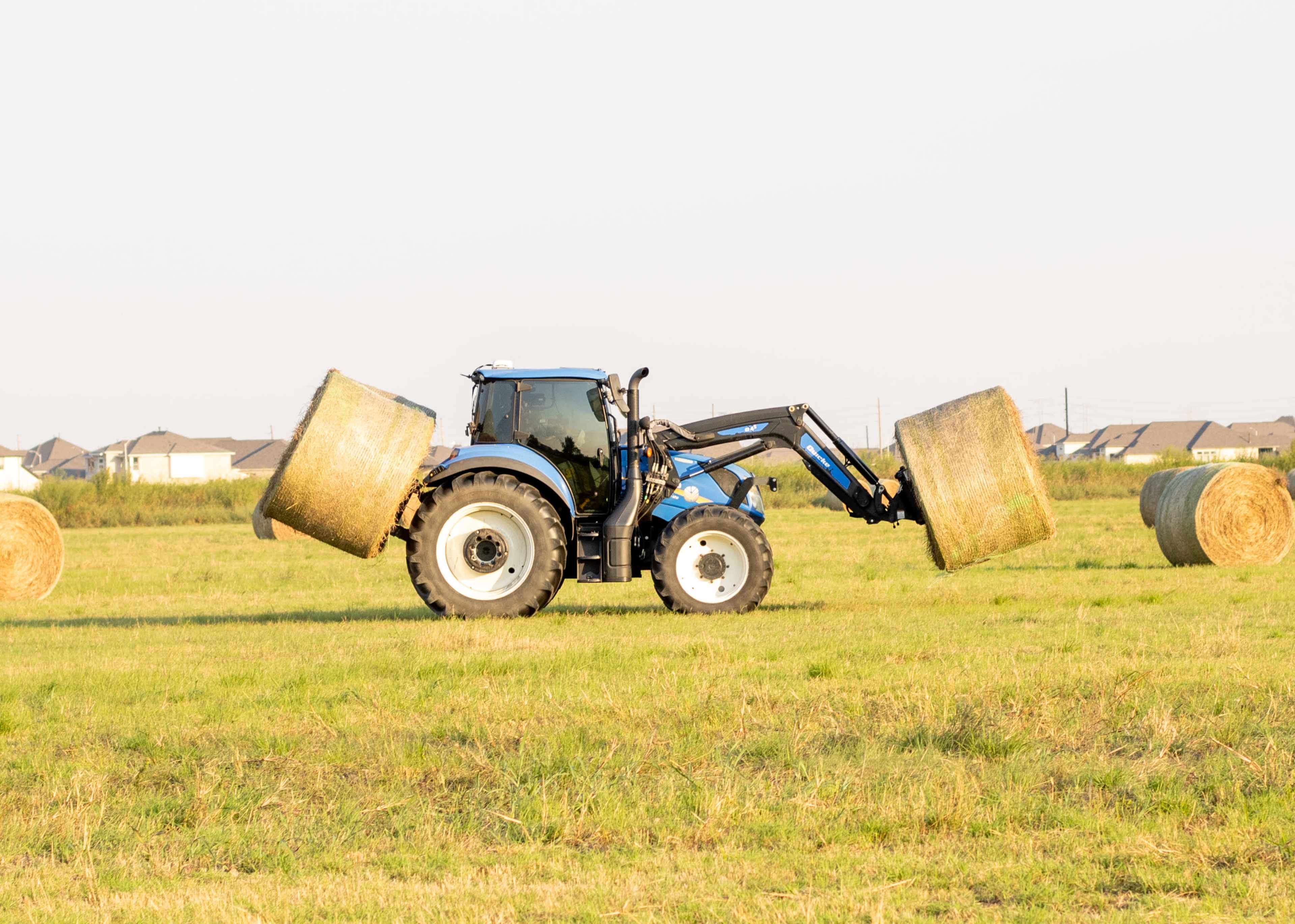 A tractor lifts large hay bales in a grassy field with houses visible in the background.
