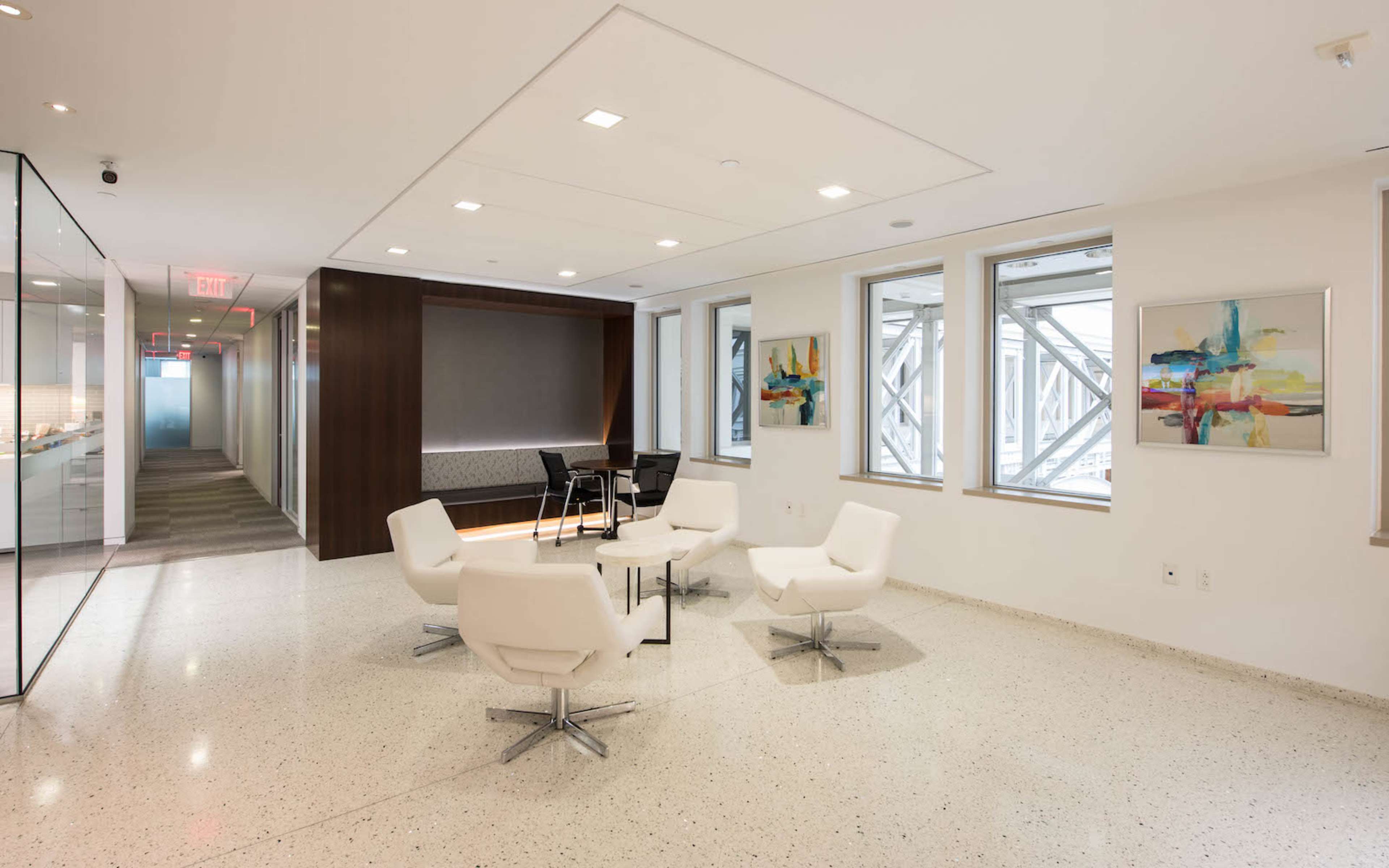 A modern office space featuring a seating area with white chairs and a glass partition, complemented by abstract artwork on the walls.
