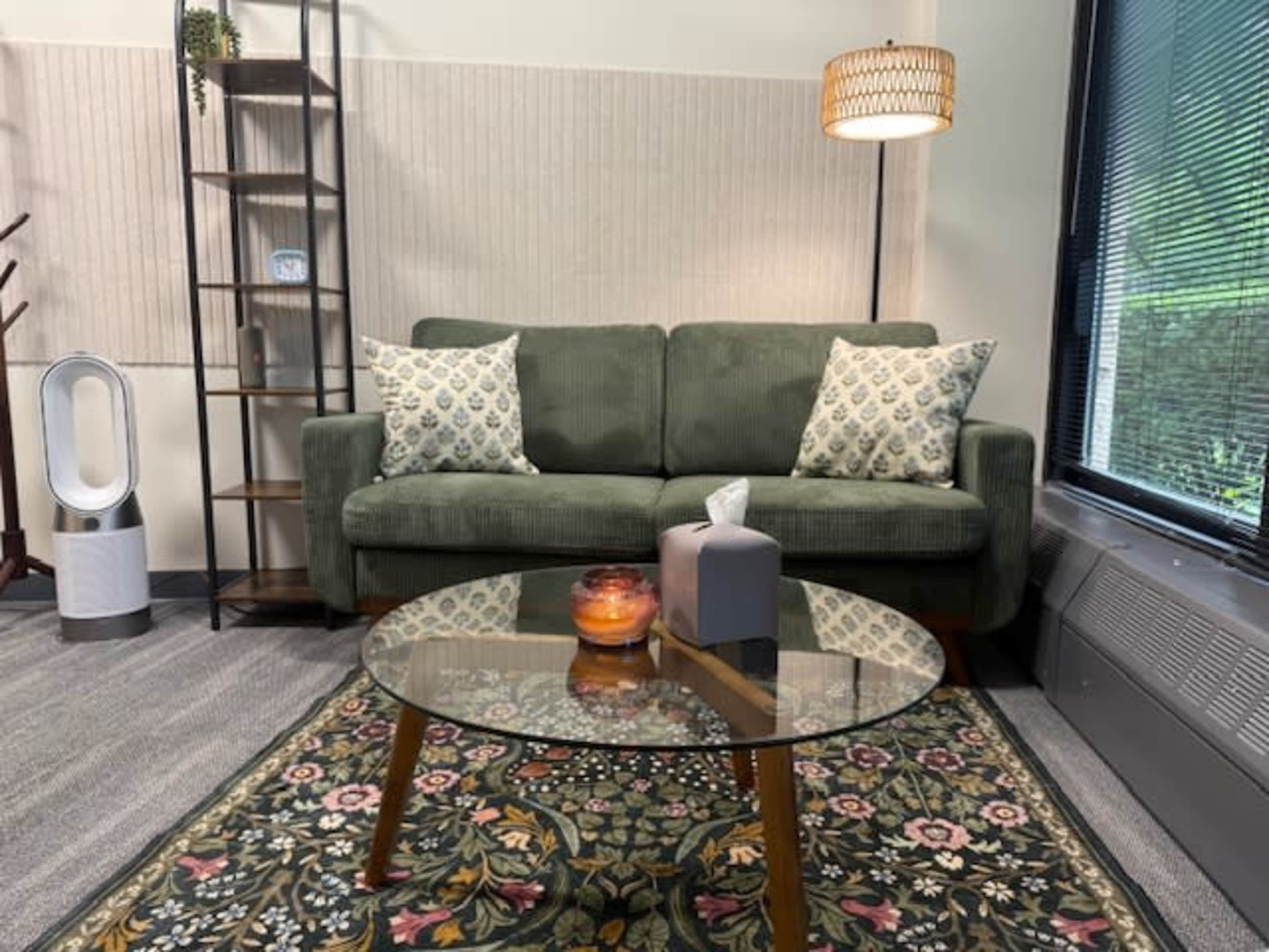 The image shows a cozy sitting area with a green couch, a round glass coffee table, decorative pillows, and a small lamp, surrounded by a patterned rug and a shelving unit.