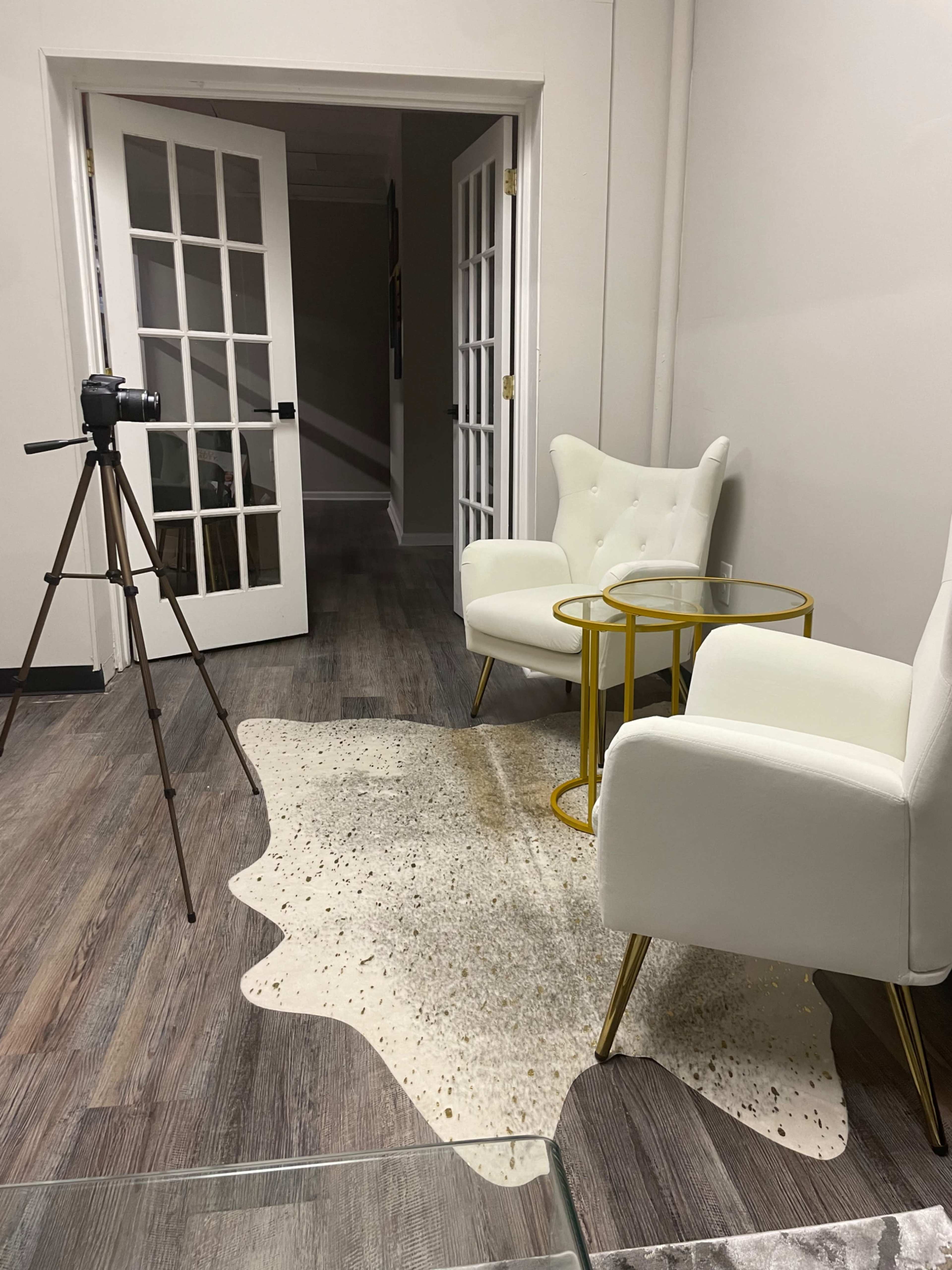 A minimalist room with two white chairs, a small gold side table, a camera on a tripod, and a light-colored rug on a wooden floor.