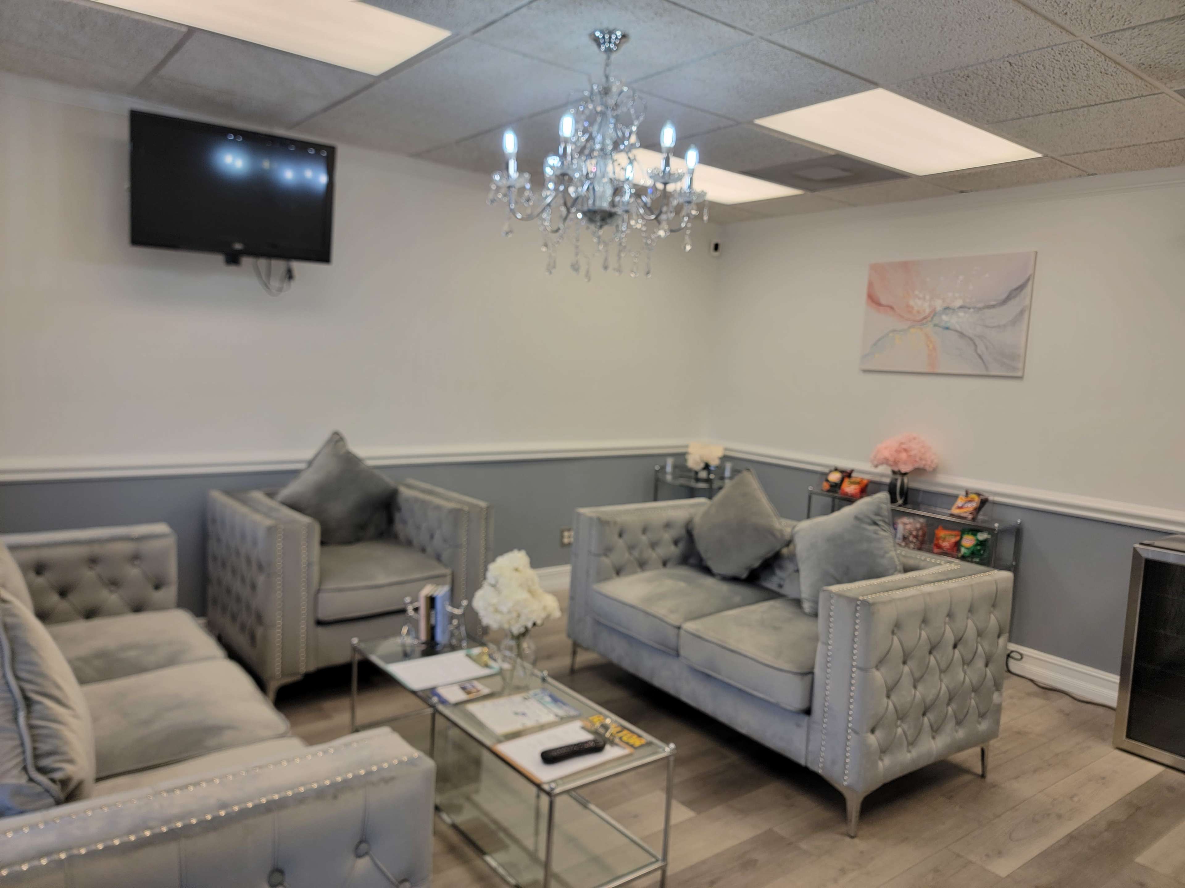 The image shows a well-furnished waiting area featuring plush gray sofas, a glass coffee table, and a chandelier, with a television mounted on the wall.