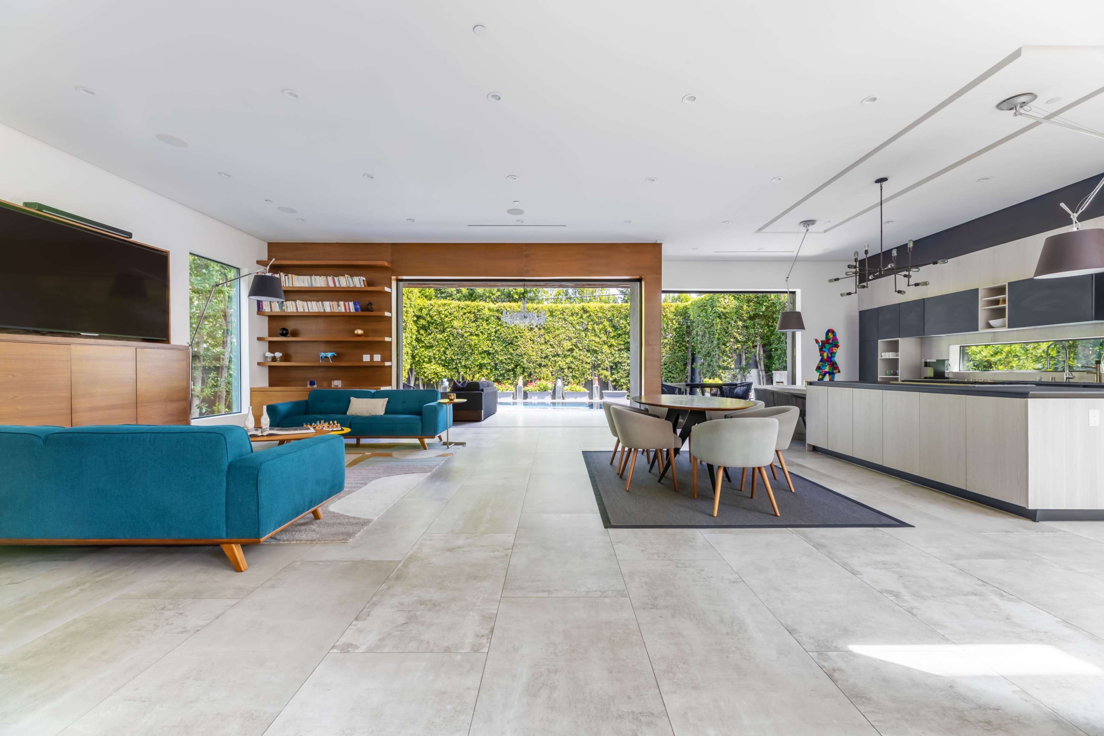 The image shows a modern living room with teal sofas, a wooden entertainment unit, and large sliding doors that open to an outdoor area.