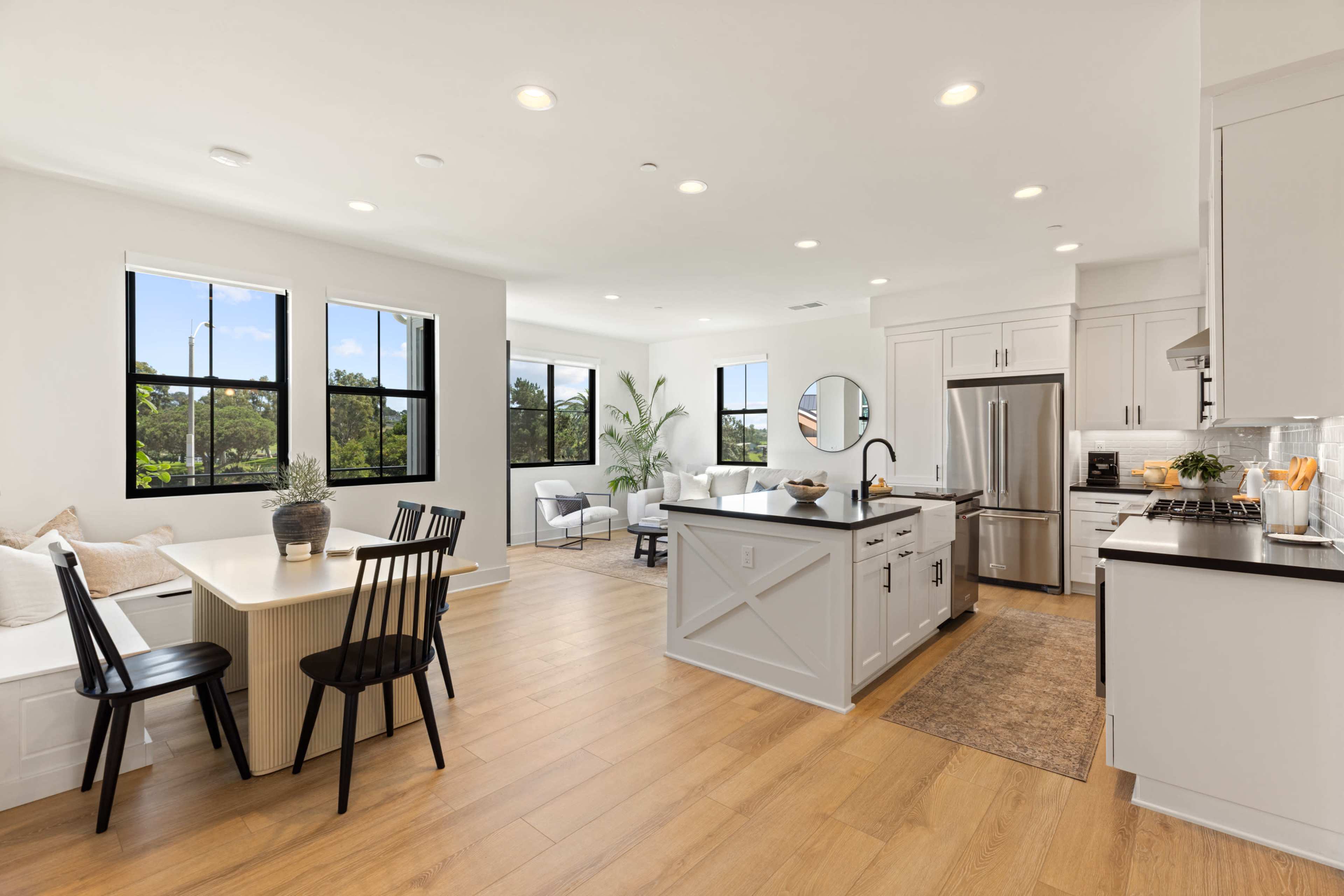 The image shows a modern kitchen and living area with large windows, a dining table, black chairs, and stainless steel appliances.