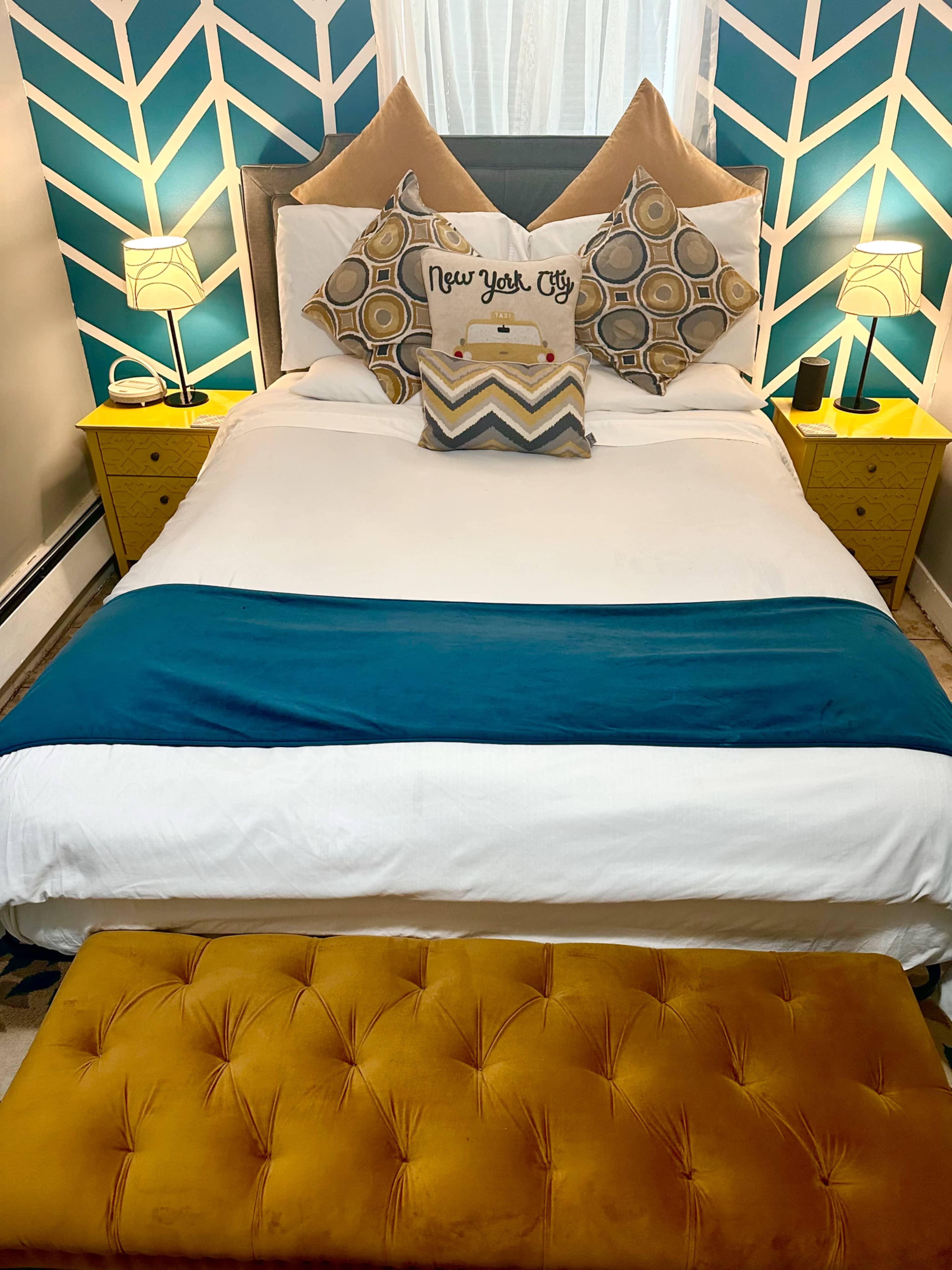 A neatly made bed with a blue accent strip, decorated with patterned pillows, flanked by two yellow nightstands and a mustard-colored bench.