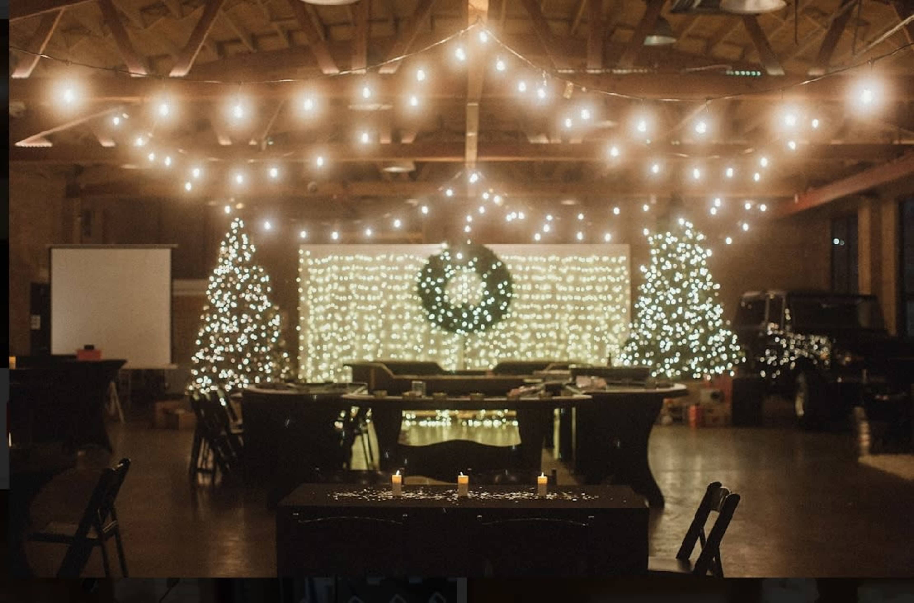 A decorated indoor space with string lights, two Christmas trees, a large wreath, and tables arranged for an event.