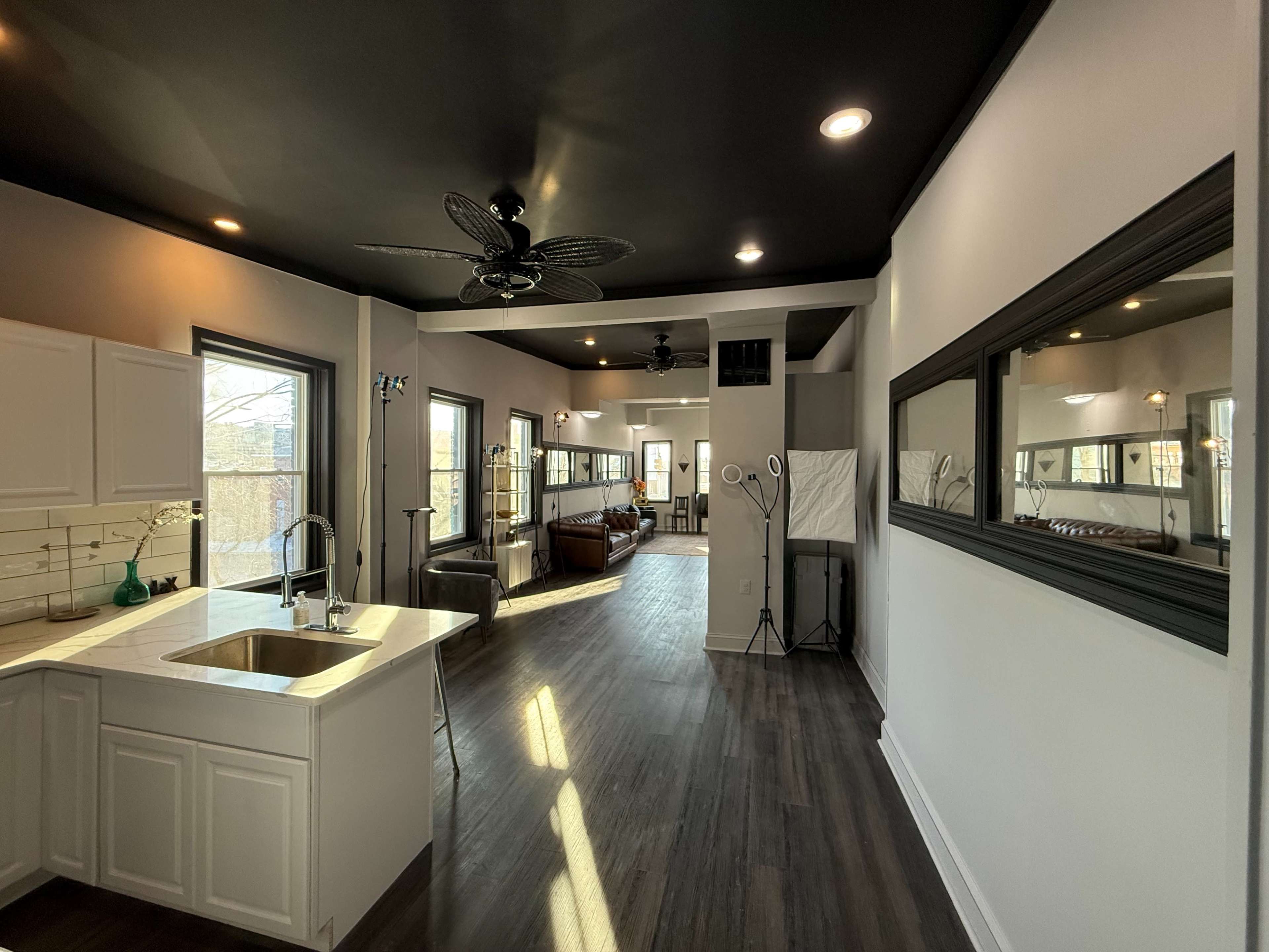 The image shows a well-lit, open-concept living space featuring a kitchen area on the left and a seating area with large windows on the right.