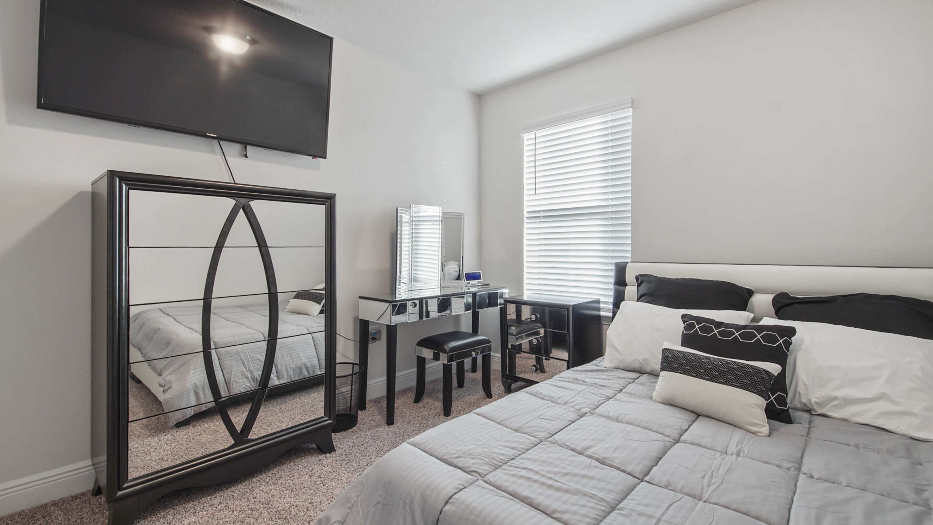 The image shows a neatly arranged bedroom featuring a bed with gray bedding, a mirrored dresser, a black vanity table with a chair, and a television mounted on the wall.