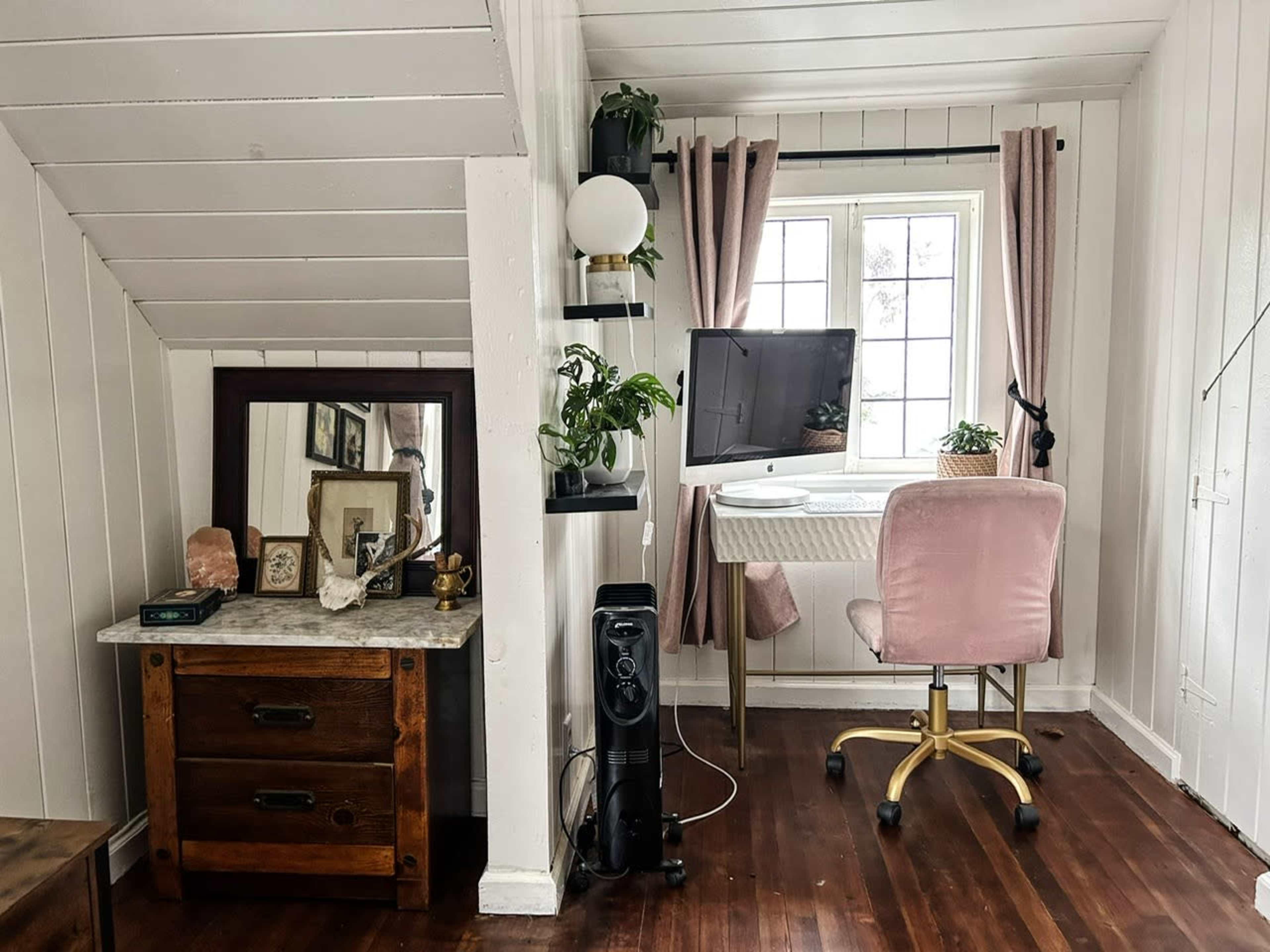 A compact home office features a desk with a computer, a pink chair, a mirror on a side table, and potted plants near a window with curtains.