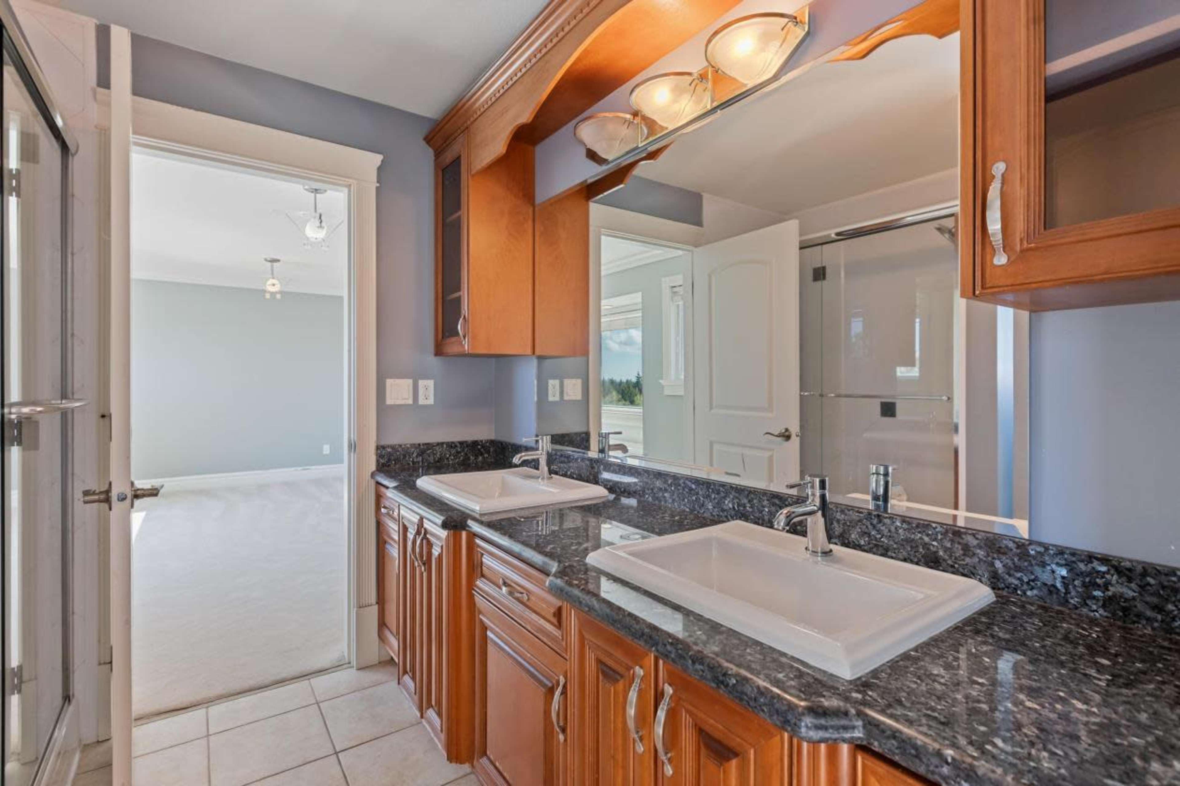 A bathroom with dual sinks, wooden cabinetry, and a large mirror, leading to a well-lit room in the background.