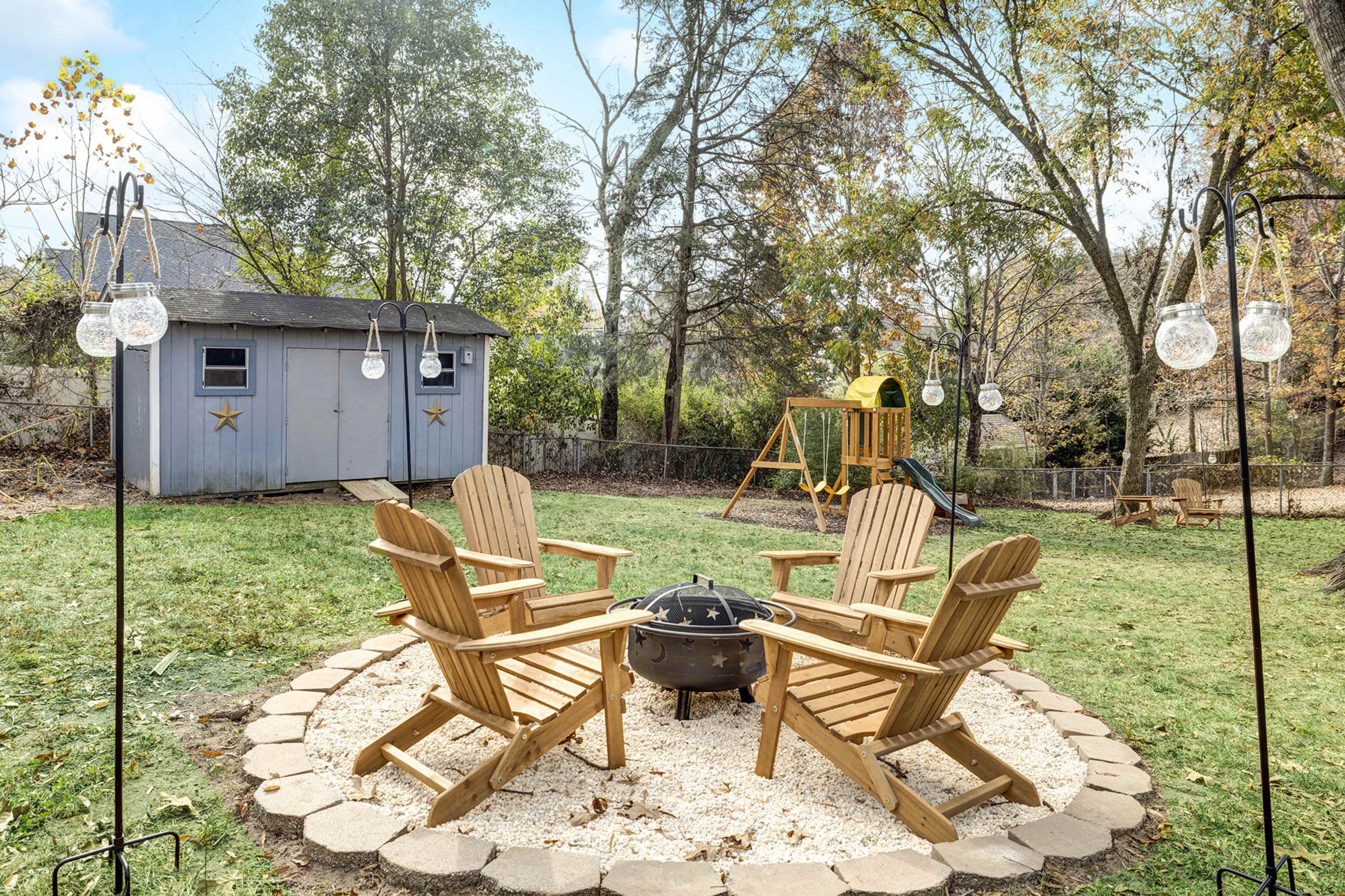 A backyard with four wooden chairs arranged around a circular fire pit on a stone patio, with a swing set and a small shed in the background.