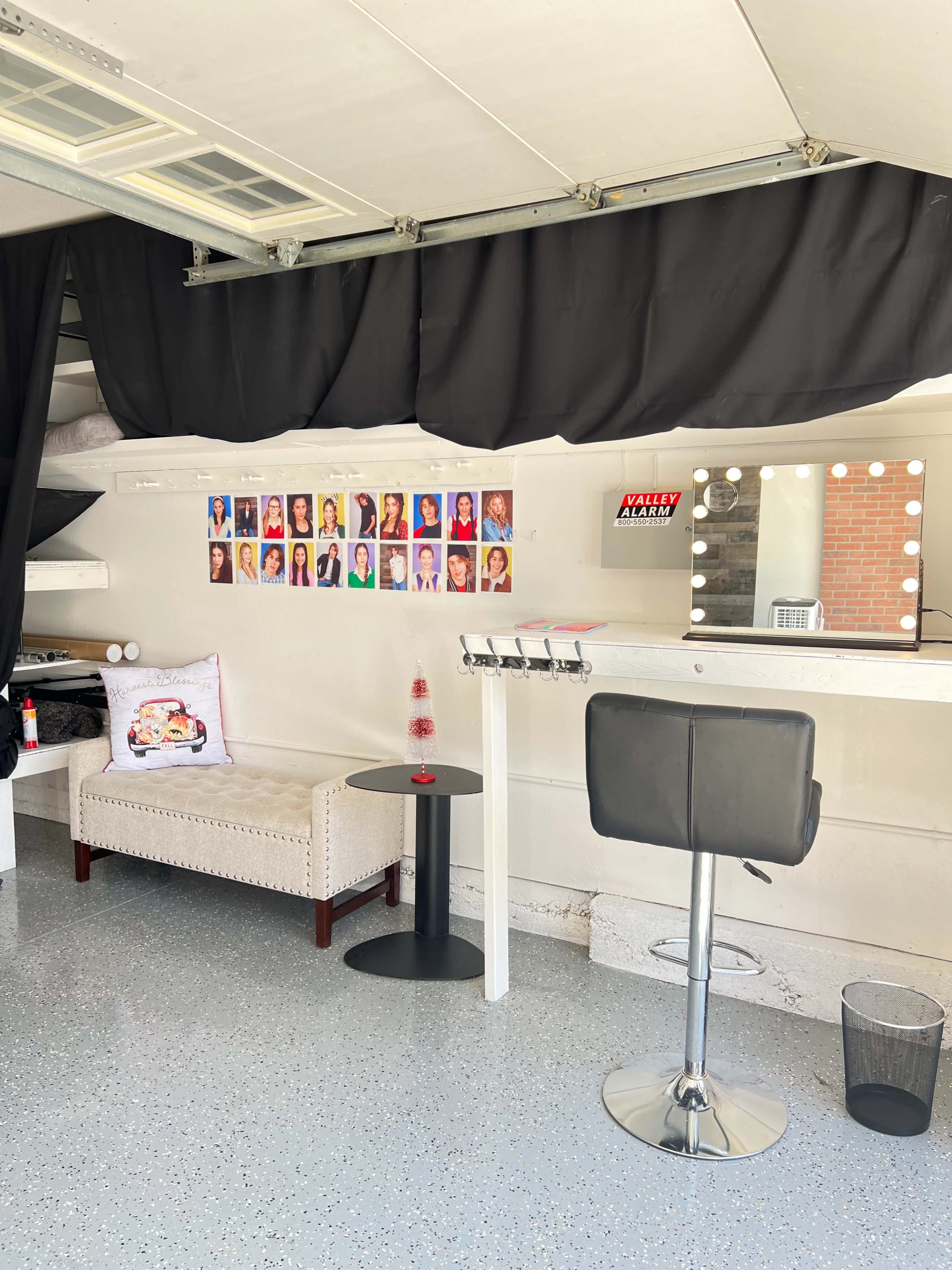 A minimalistic beauty salon interior featuring a couch, a bar stool, a round table, a wall of portraits, and a well-lit makeup mirror.