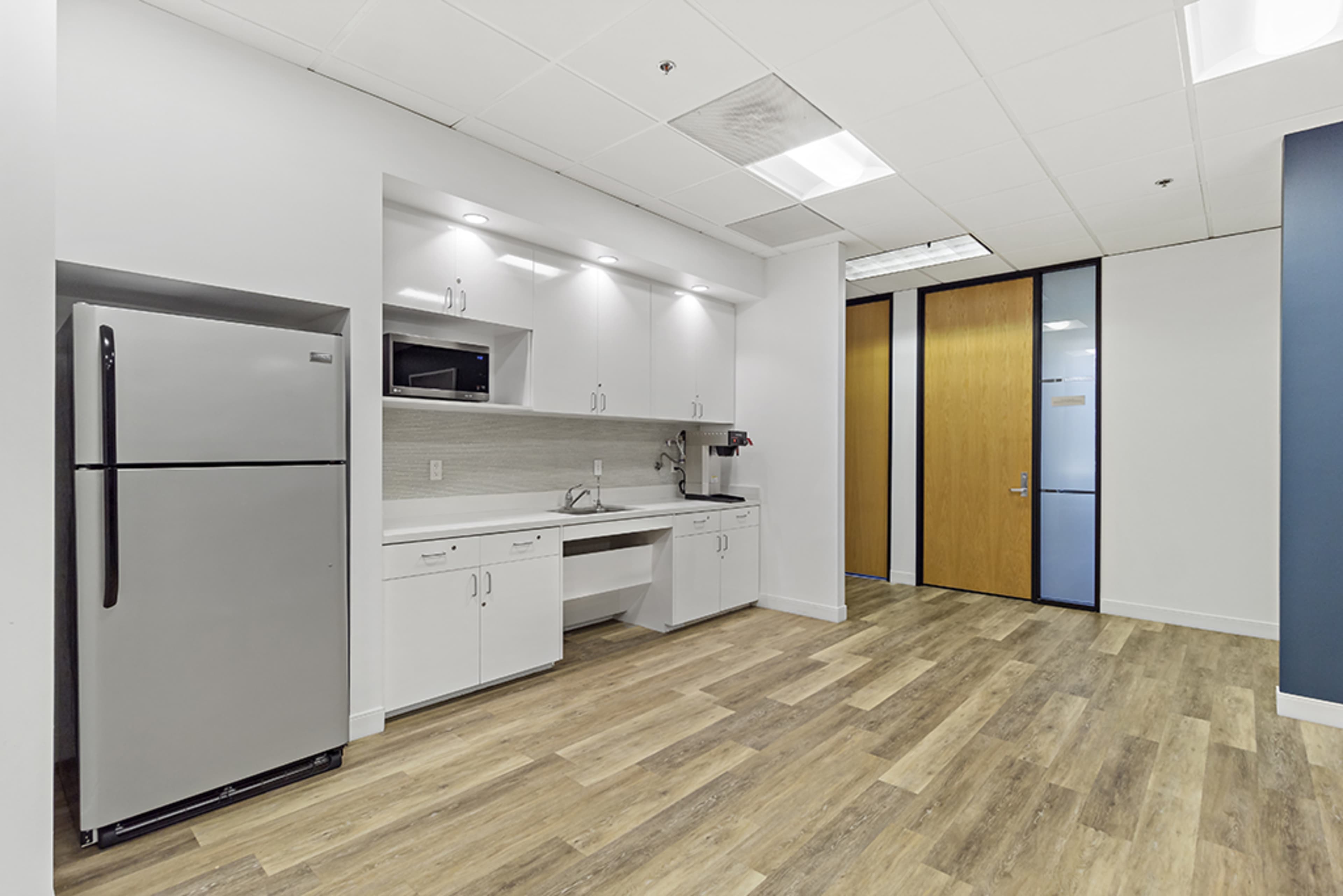 A modern kitchen area featuring a stainless steel refrigerator, a microwave above the counter, and a sink, with laminate flooring and glass-panel doors leading to another room.