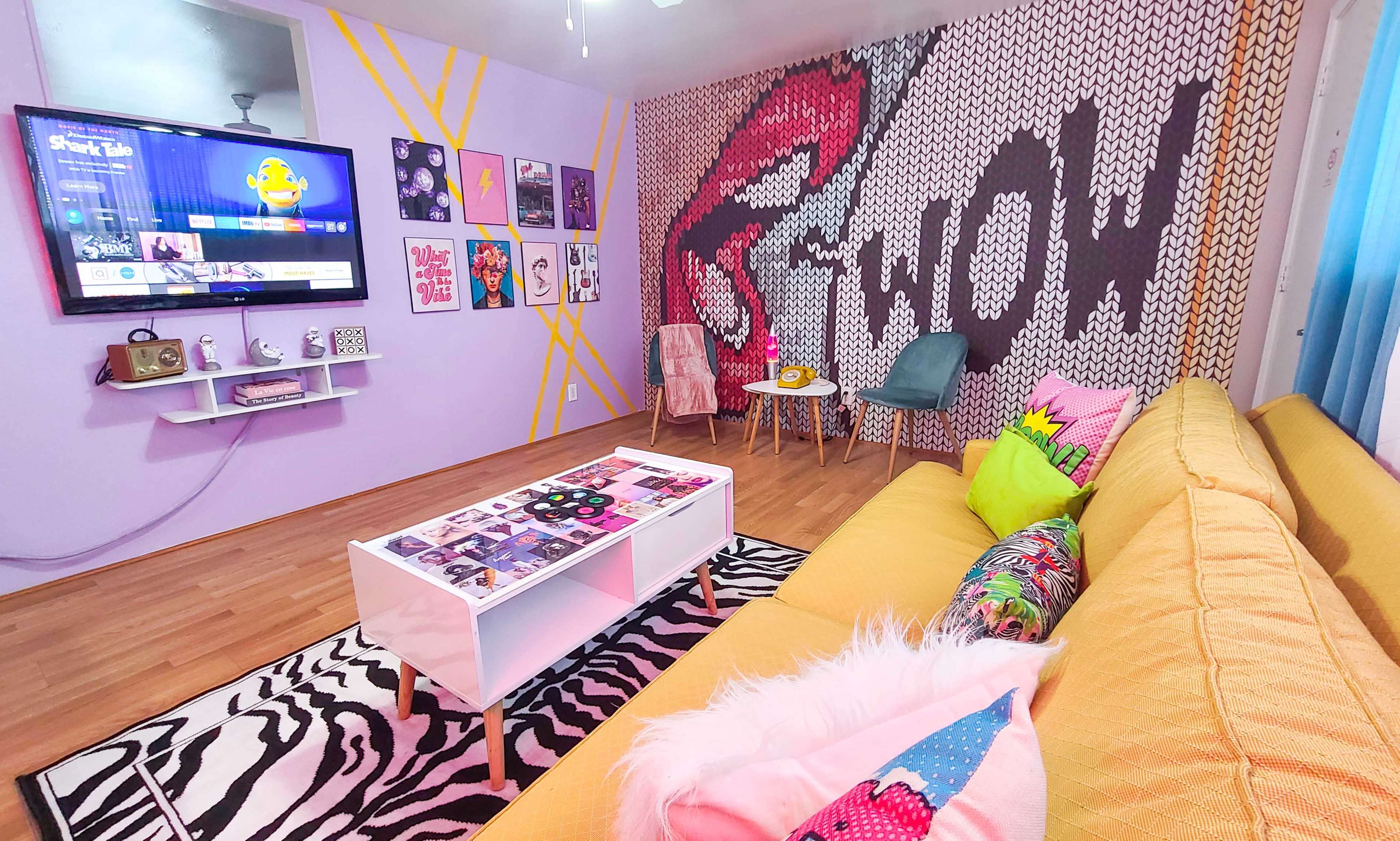 The image shows a colorful living room with a yellow sofa, a coffee table covered in magazines, and a wall featuring vibrant pop art and a television display.