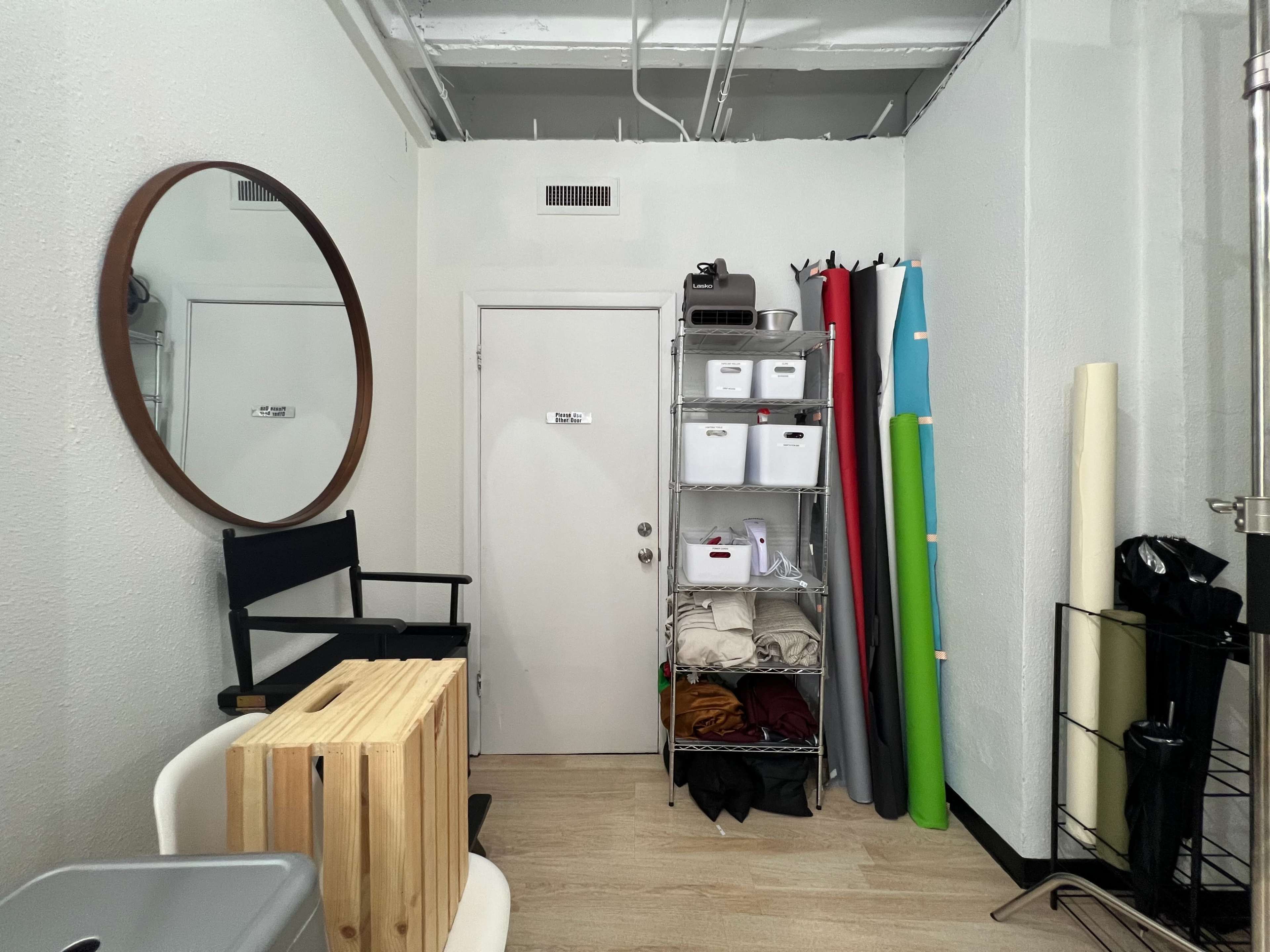 A small studio room with a round mirror, a door, a shelving unit filled with storage boxes, and rolls of colored paper stacked against the wall.