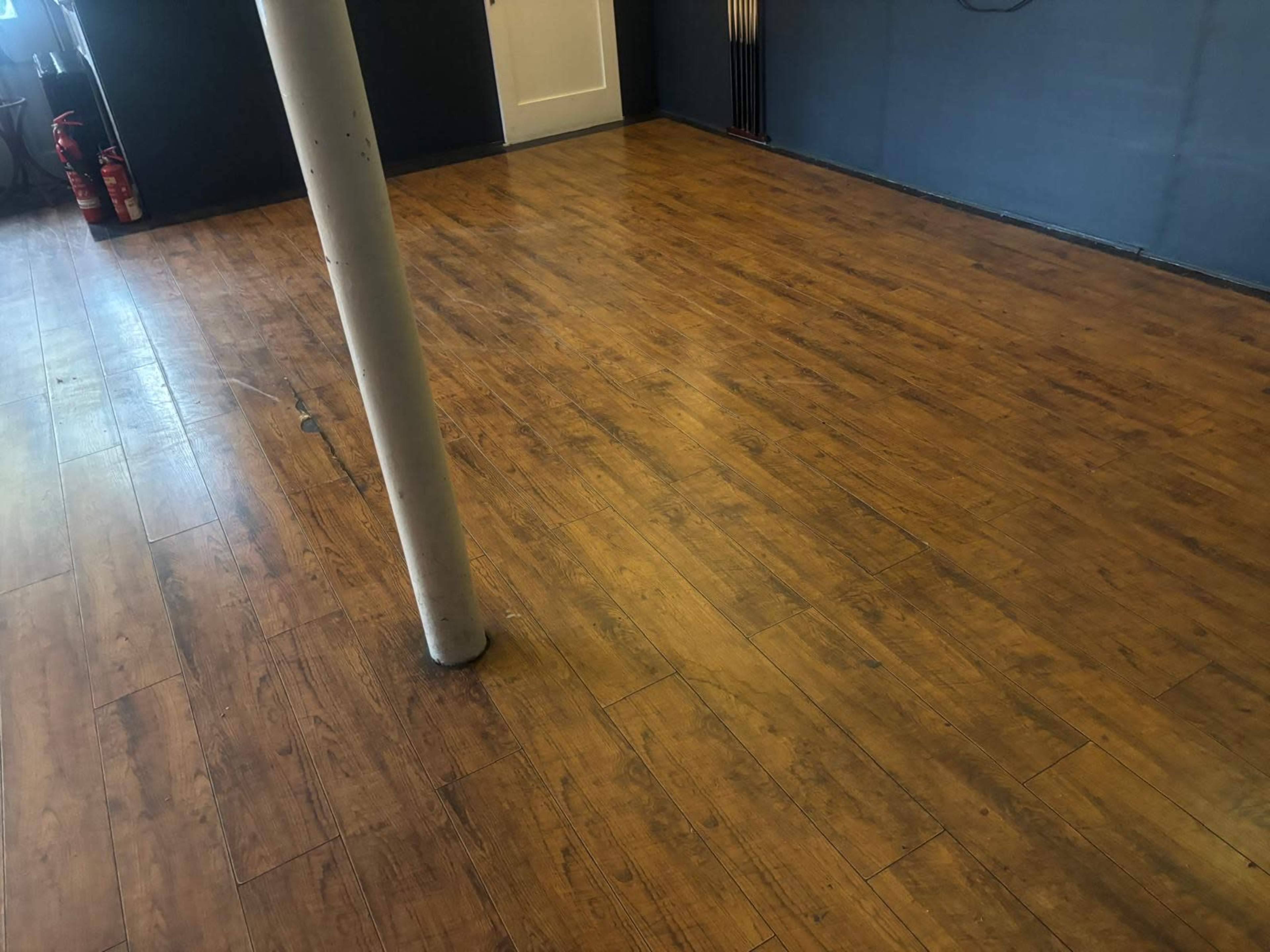 The image shows a wooden floor with a single vertical support pole in a room that has a plain wall and a door in the background.