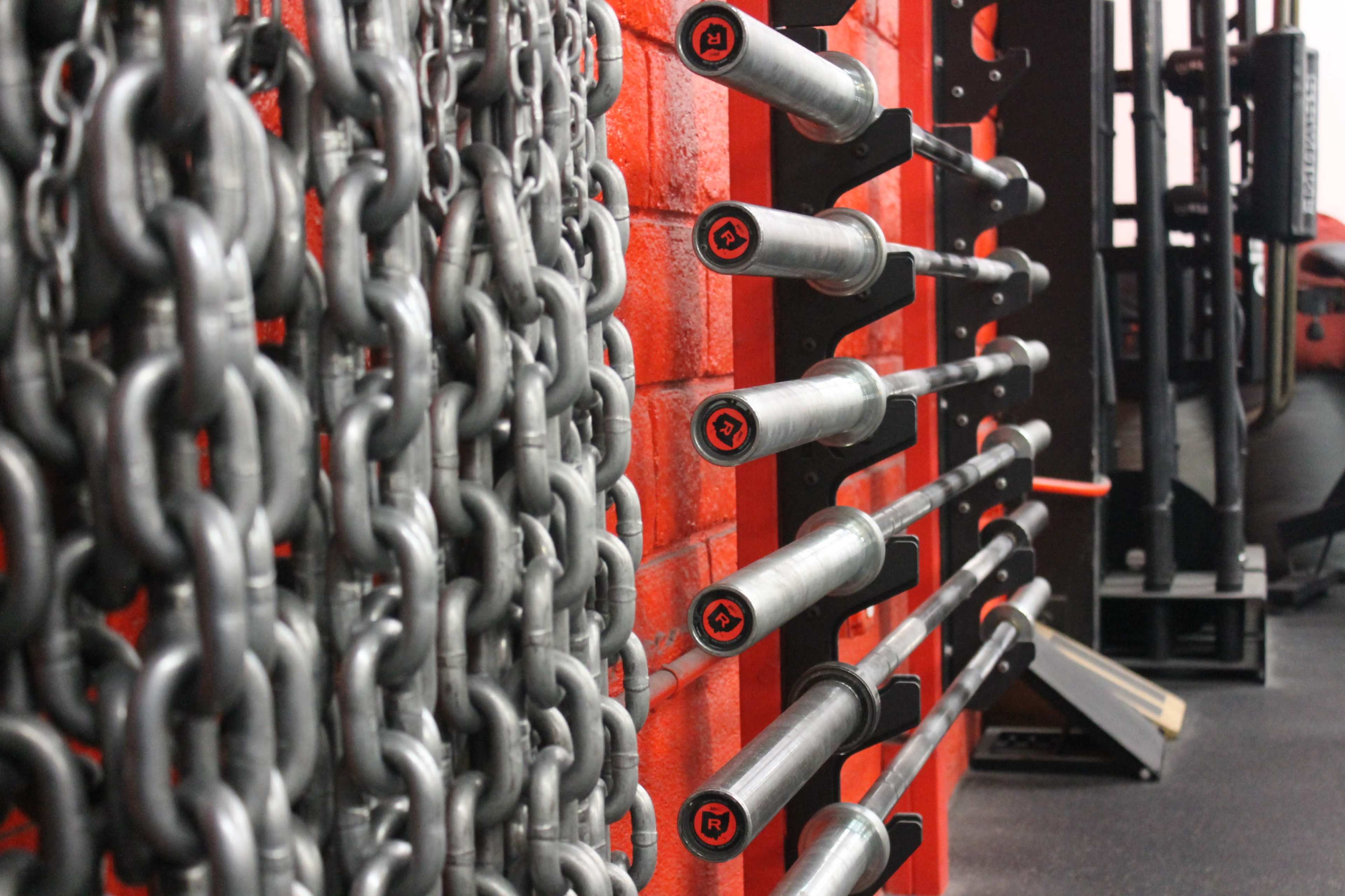 A gym interior with an orange wall featuring metal weightlifting bars mounted on it and chains hanging nearby.