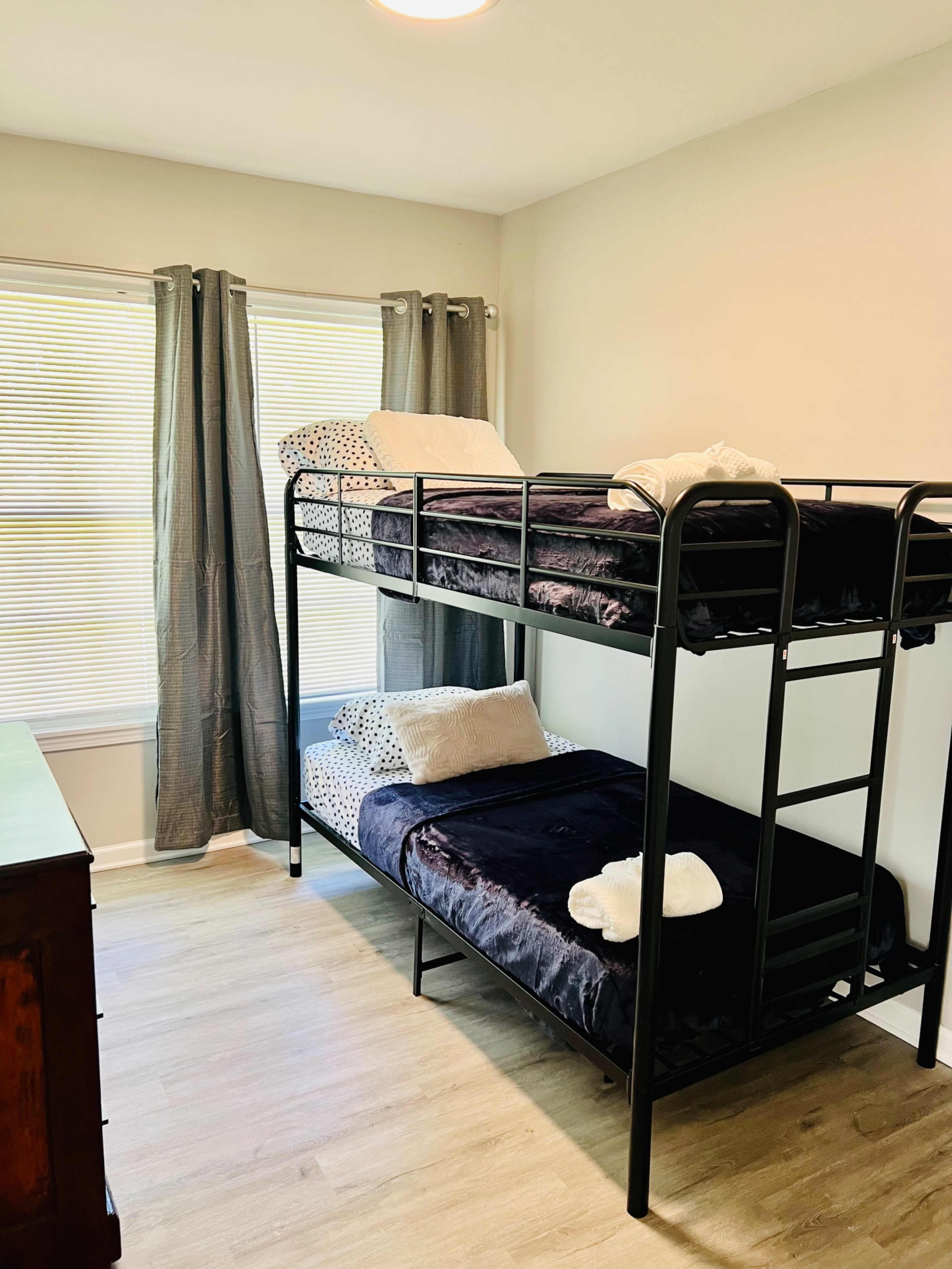 A room with a black metal bunk bed, two neatly made beds with dark bedding, and a dresser under a window with light gray curtains.