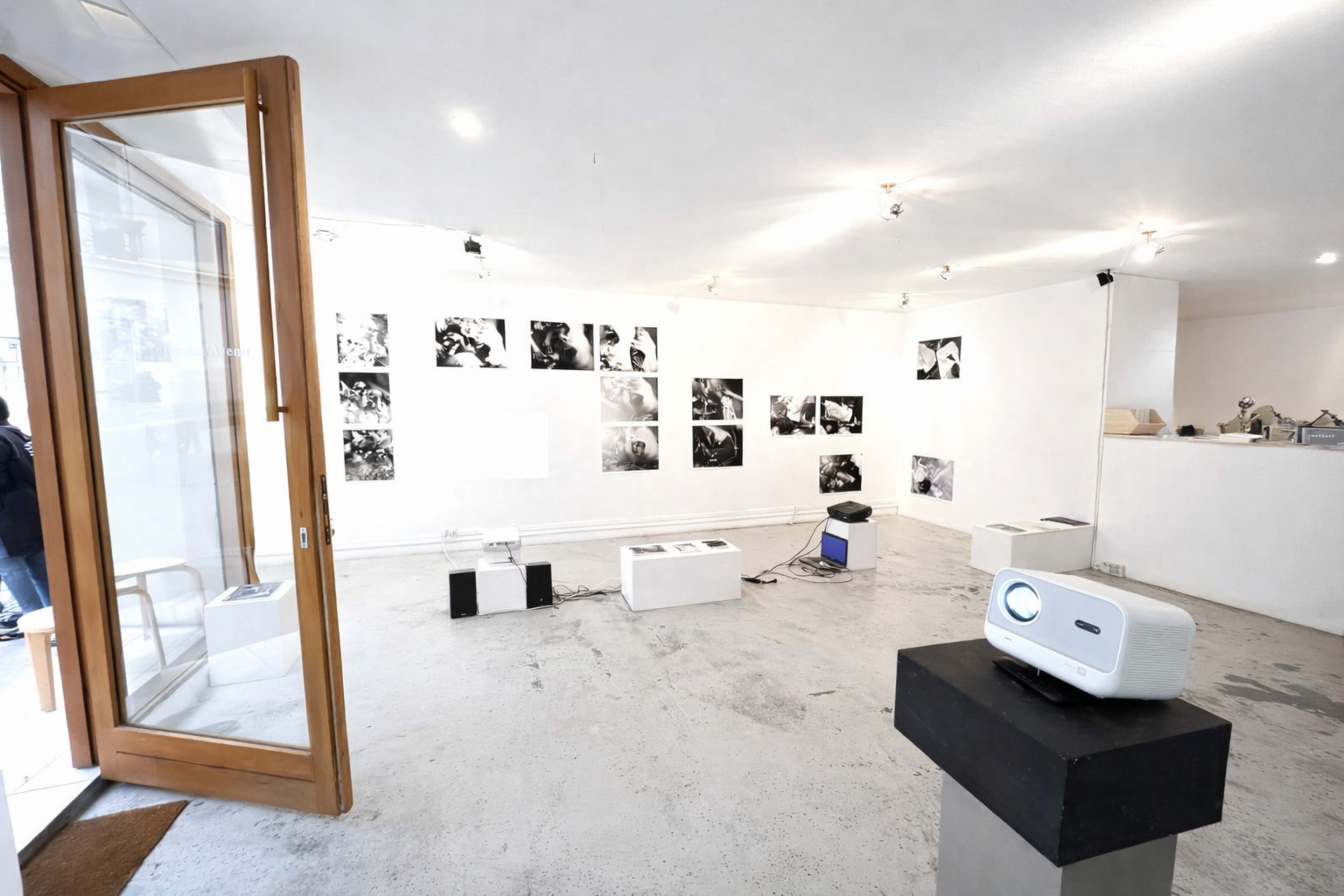 The image shows an art gallery featuring black and white photographs on the walls and a projector set up on a stand in the foreground.