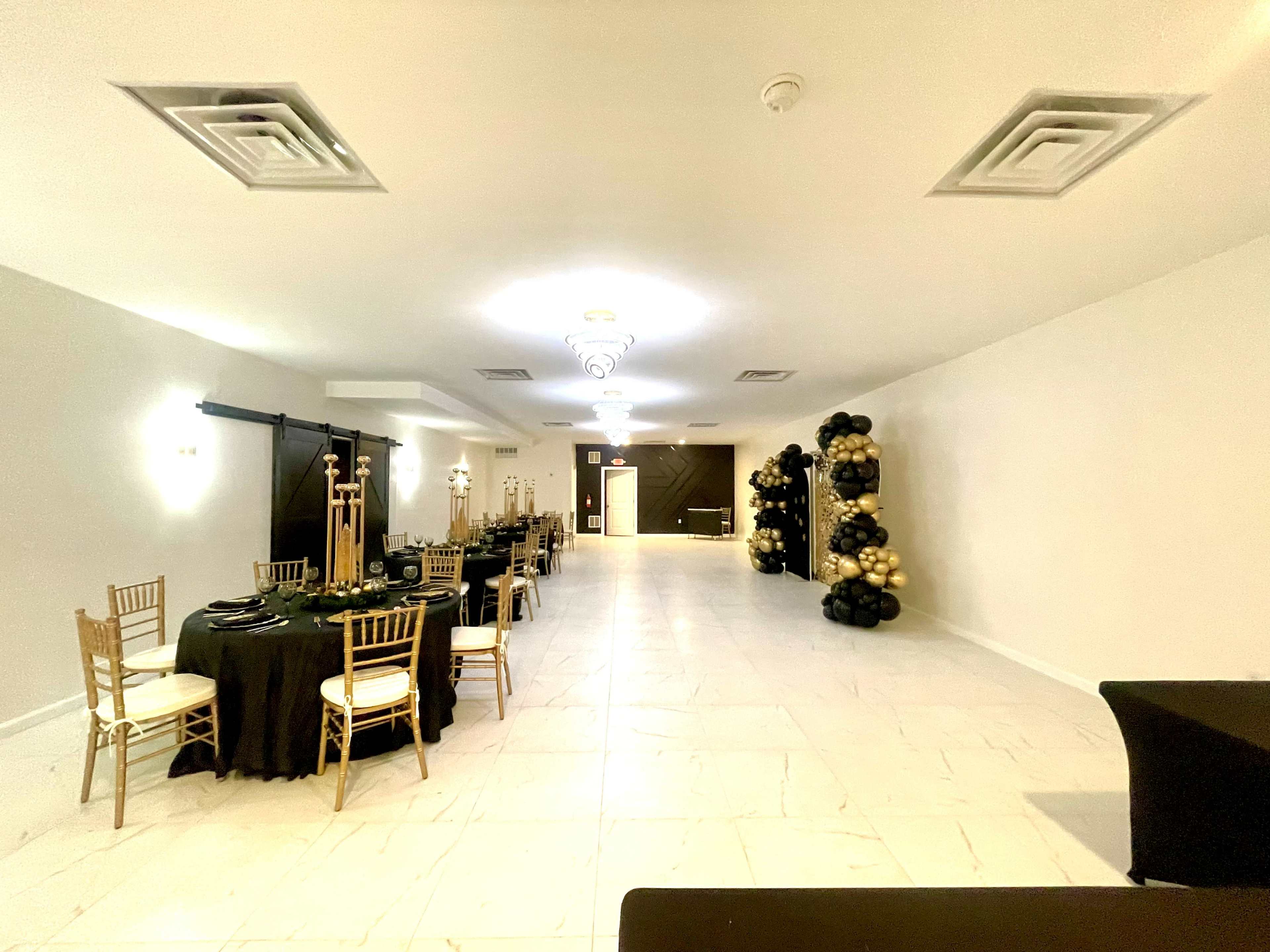 A spacious event hall is set with black and gold table settings, featuring gold chiavari chairs and a decorative arrangement of black and gold balloons at one end.
