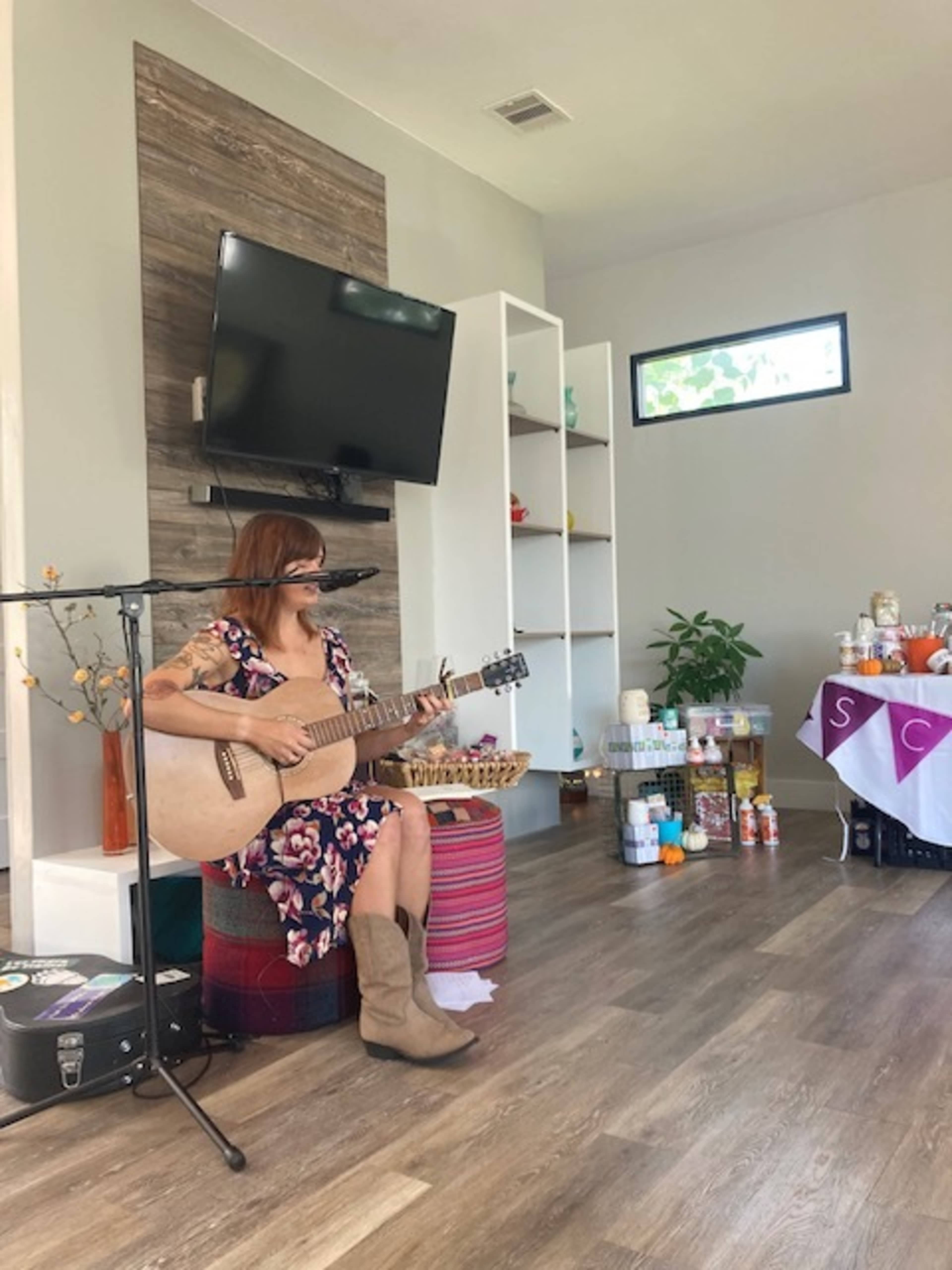 A woman plays guitar and sings on a stool in a brightly lit room with a television and shelves in the background.