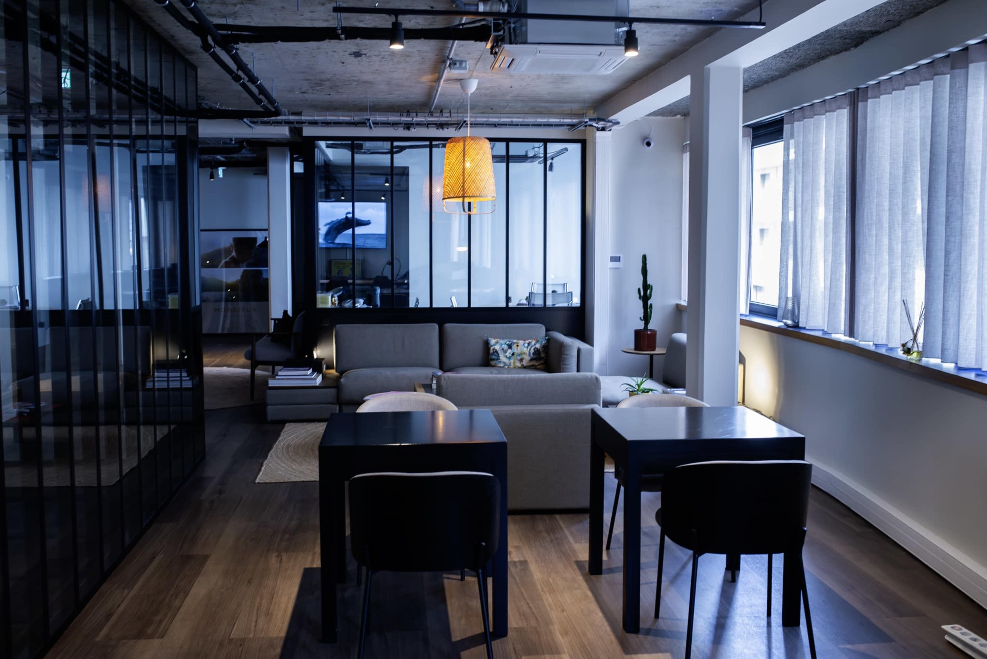 The image shows a modern open office space with a seating area featuring a sofa, a table for two, and large windows allowing natural light.