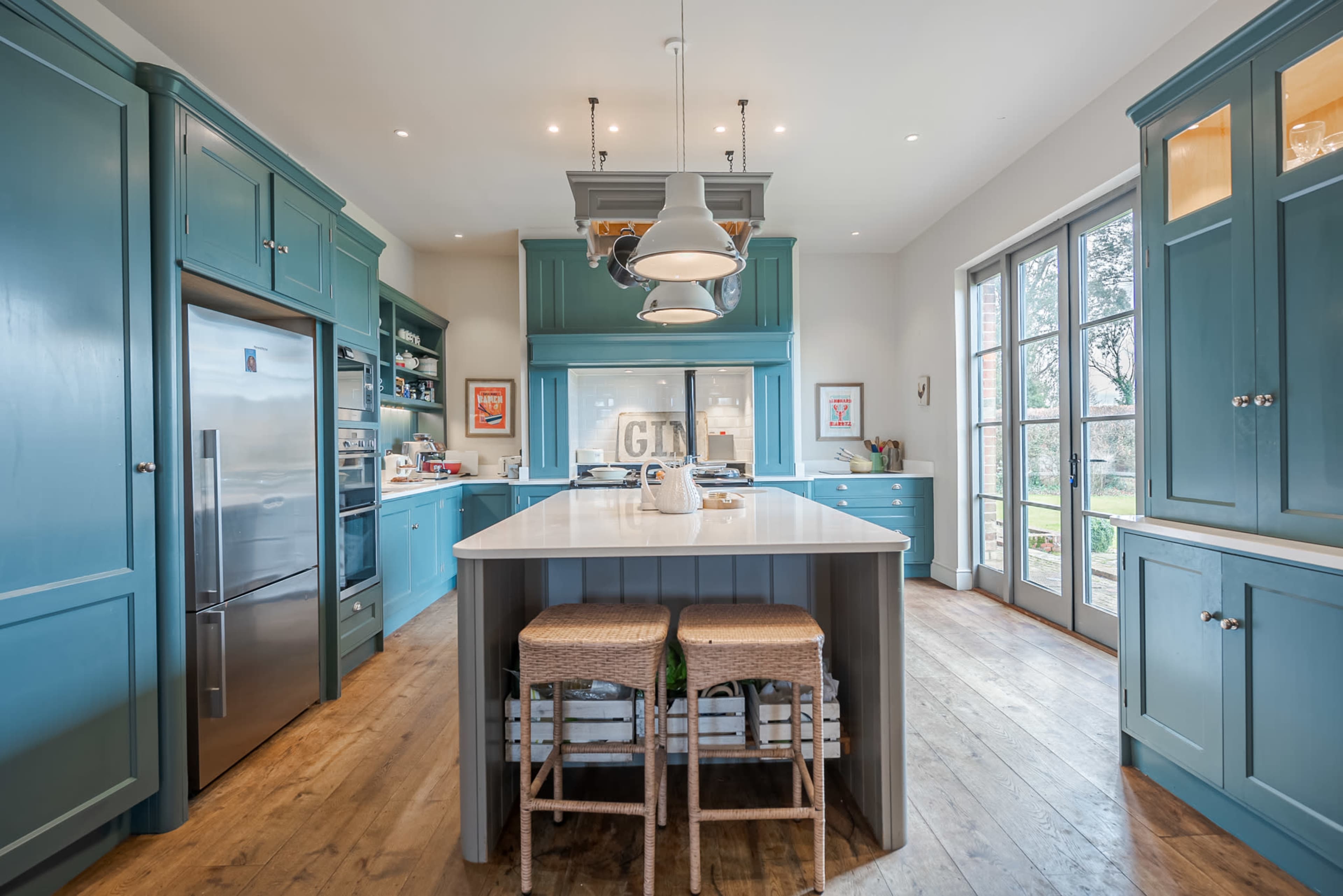 A modern kitchen with blue cabinetry, a large central island with two wicker stools, and large windows allowing natural light to enter.