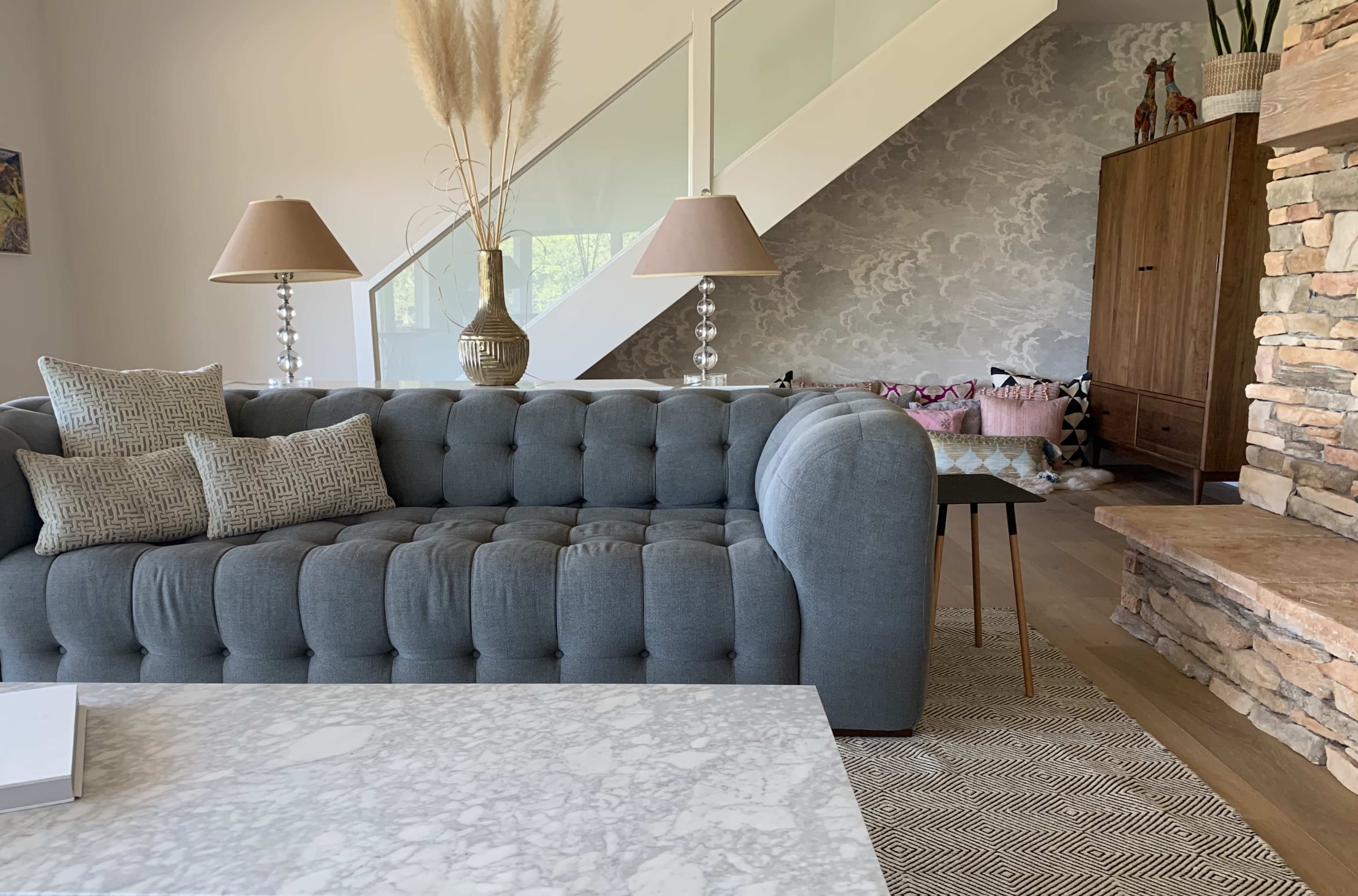 A modern living room featuring a gray tufted sofa, marble coffee table, and a staircase in the background, along with decorative lamps and a wooden cabinet.