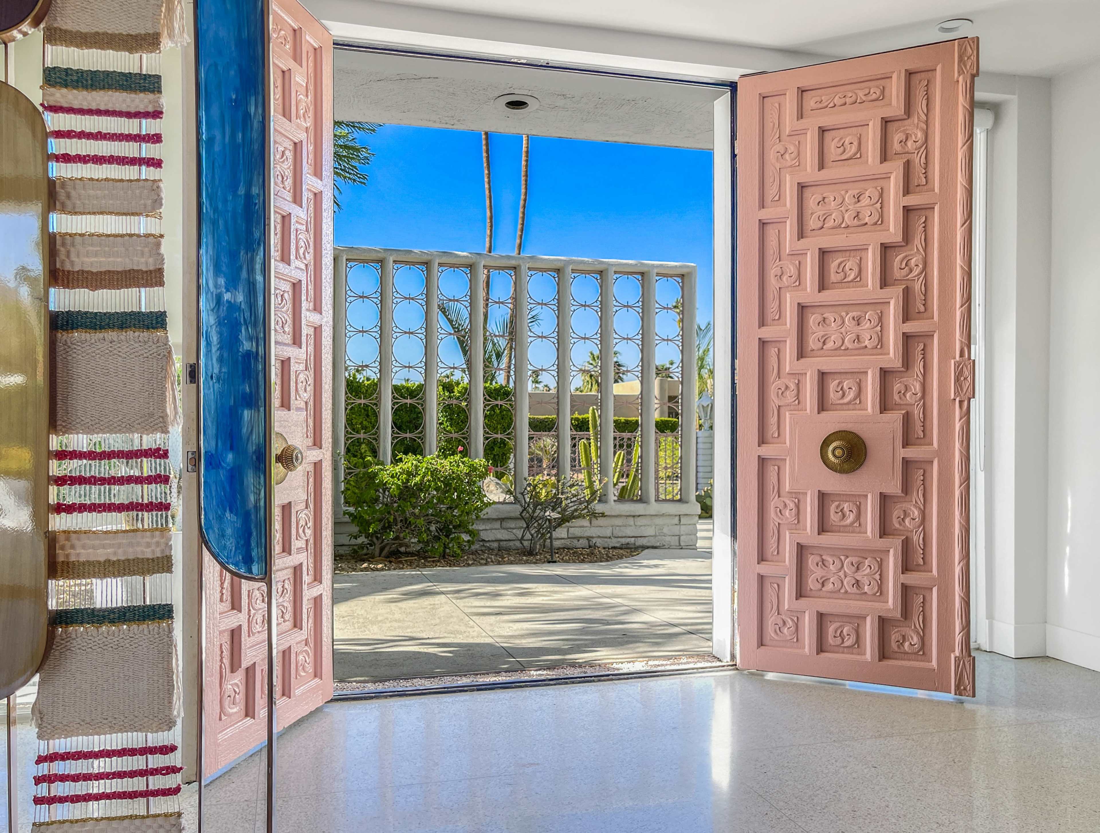 A pair of pink doors opens to a view of greenery and a decorative fence under a clear blue sky.