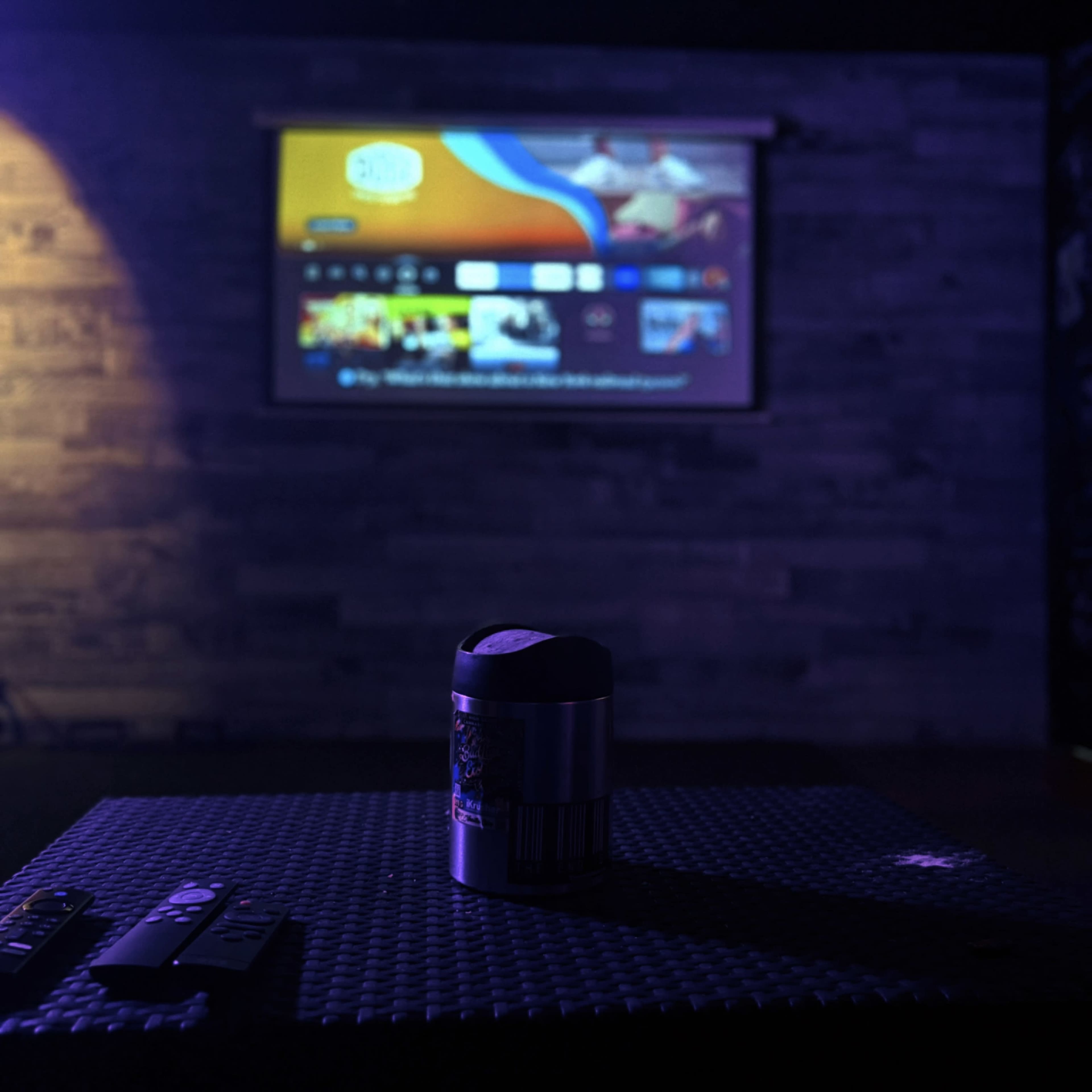 A remote control lays on a textured table in the foreground, while a projection screen displays a colorful menu in the background.