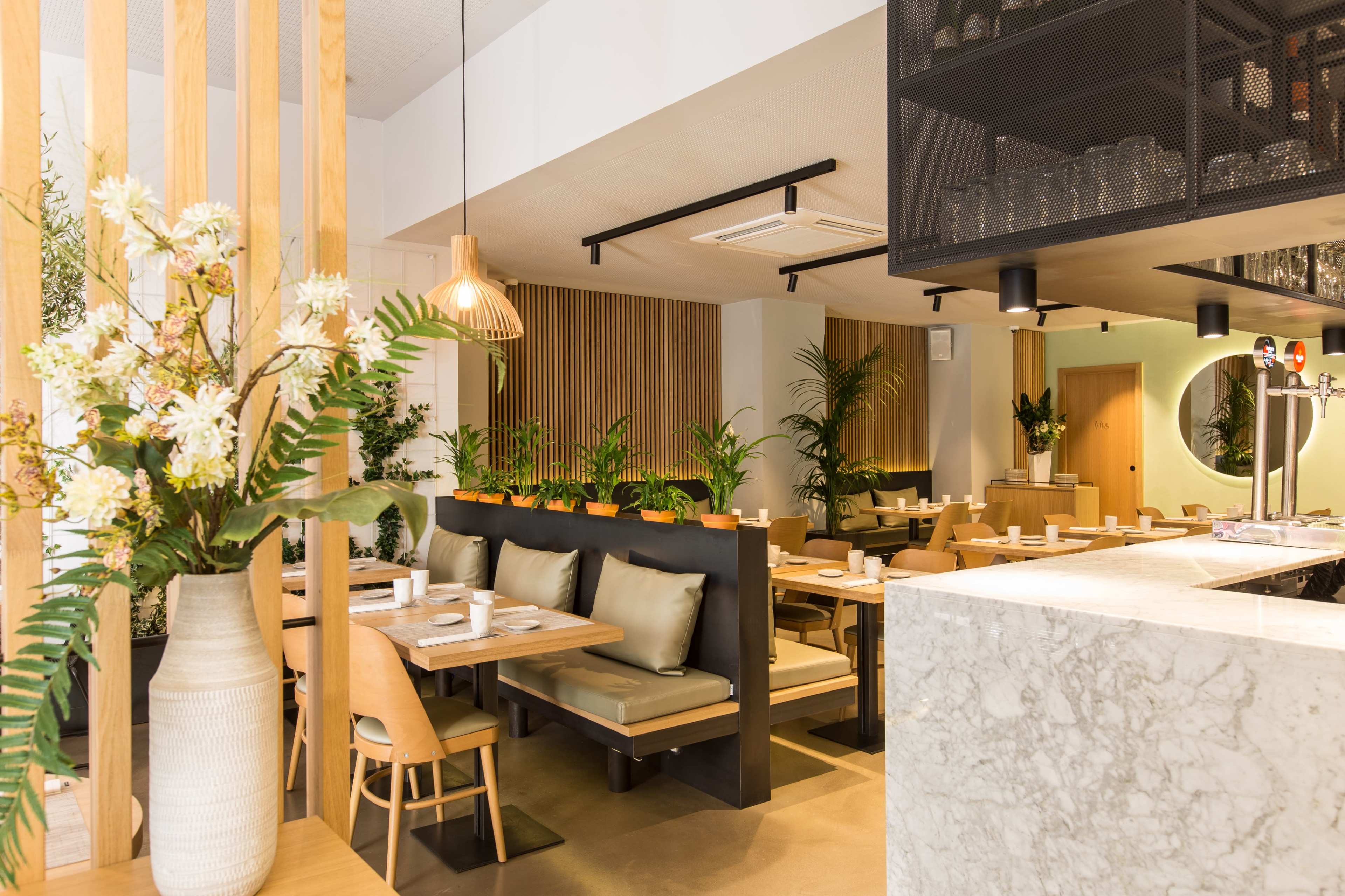 A modern restaurant interior featuring wooden accents, seating areas with cushions, and decorative plants throughout.