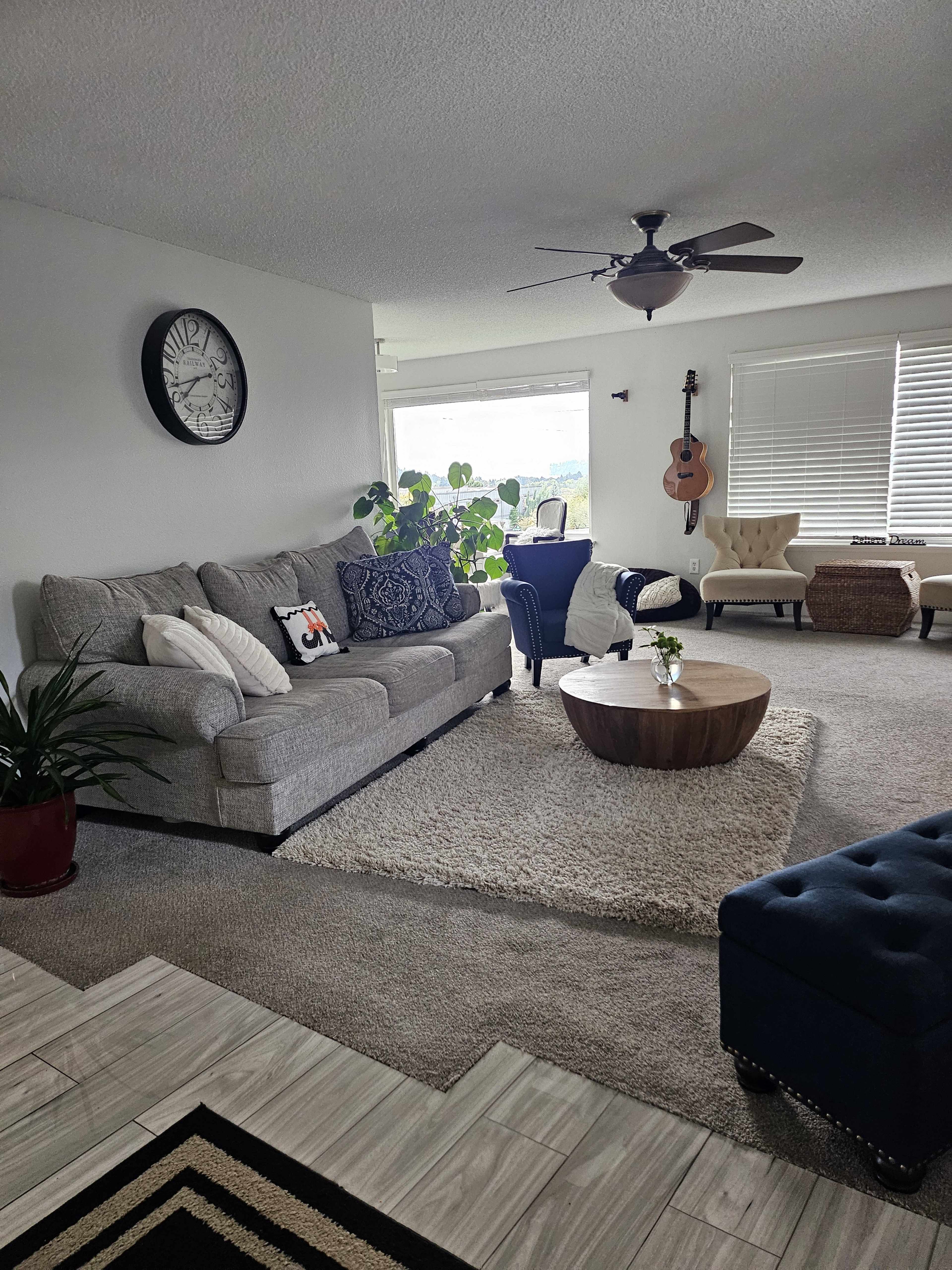 The living room features a gray sofa, a round wooden coffee table, a blue chair, and potted plants, with large windows allowing natural light.