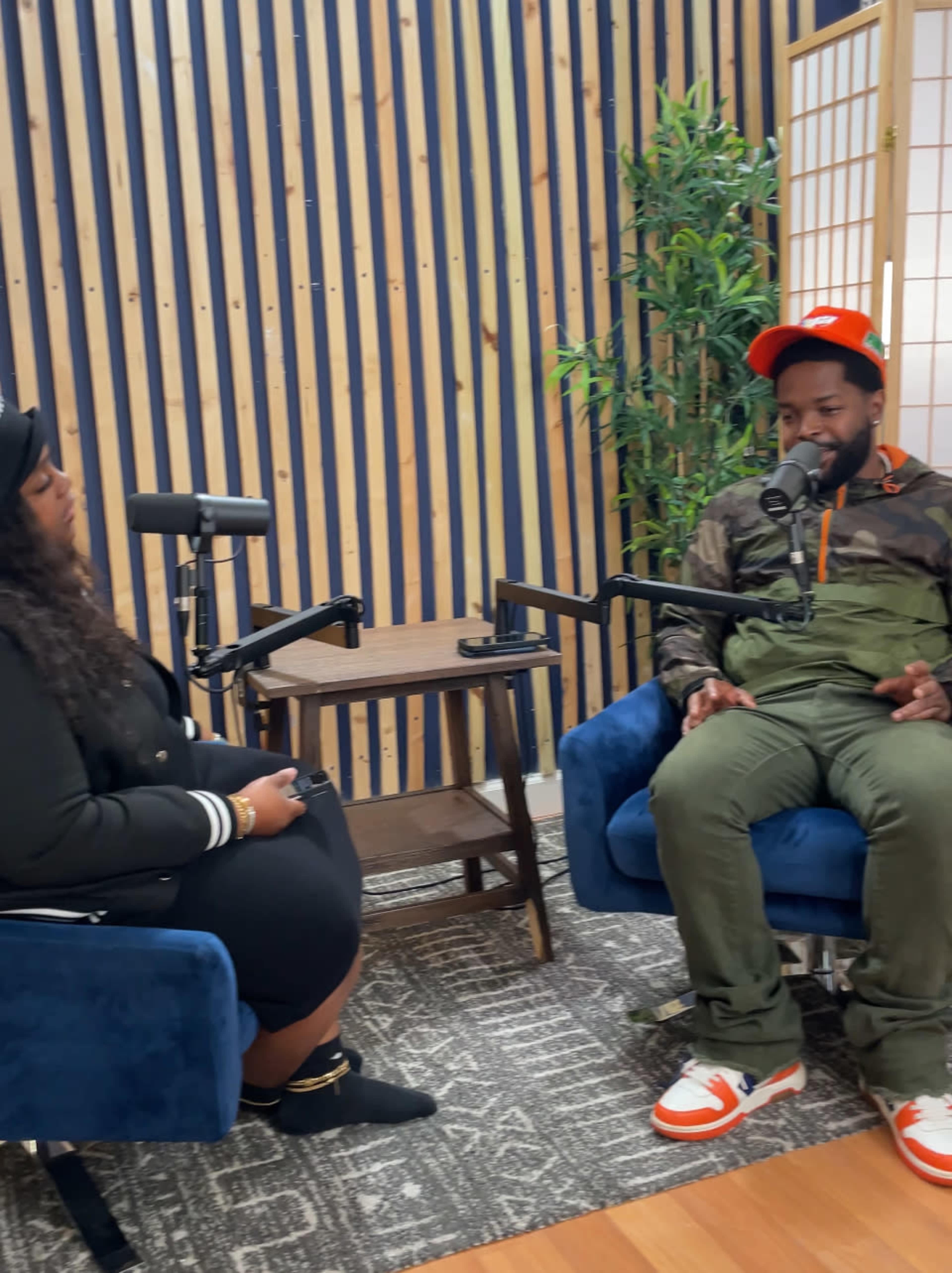 A woman and a man are seated in a modern studio with wooden paneling, engaging in conversation while microphones are positioned before them.