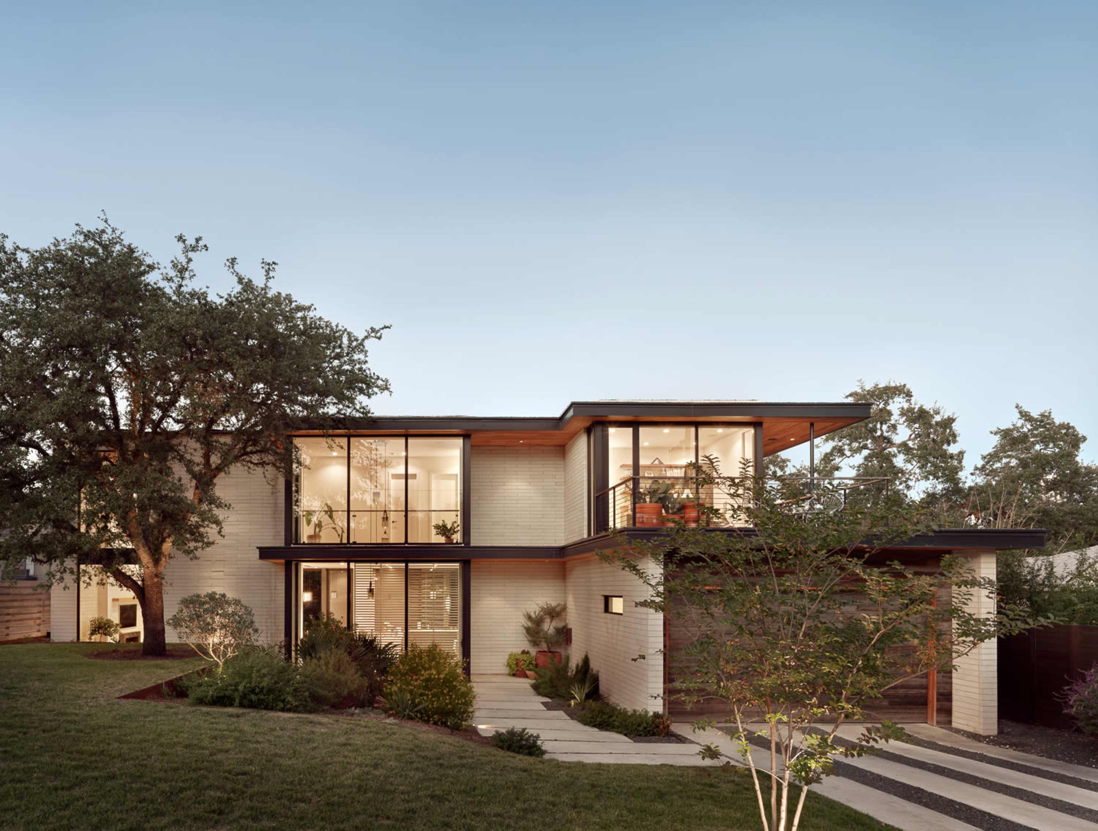 A modern two-story house features large glass windows, a wooden overhang, and landscaped greenery surrounding the property.