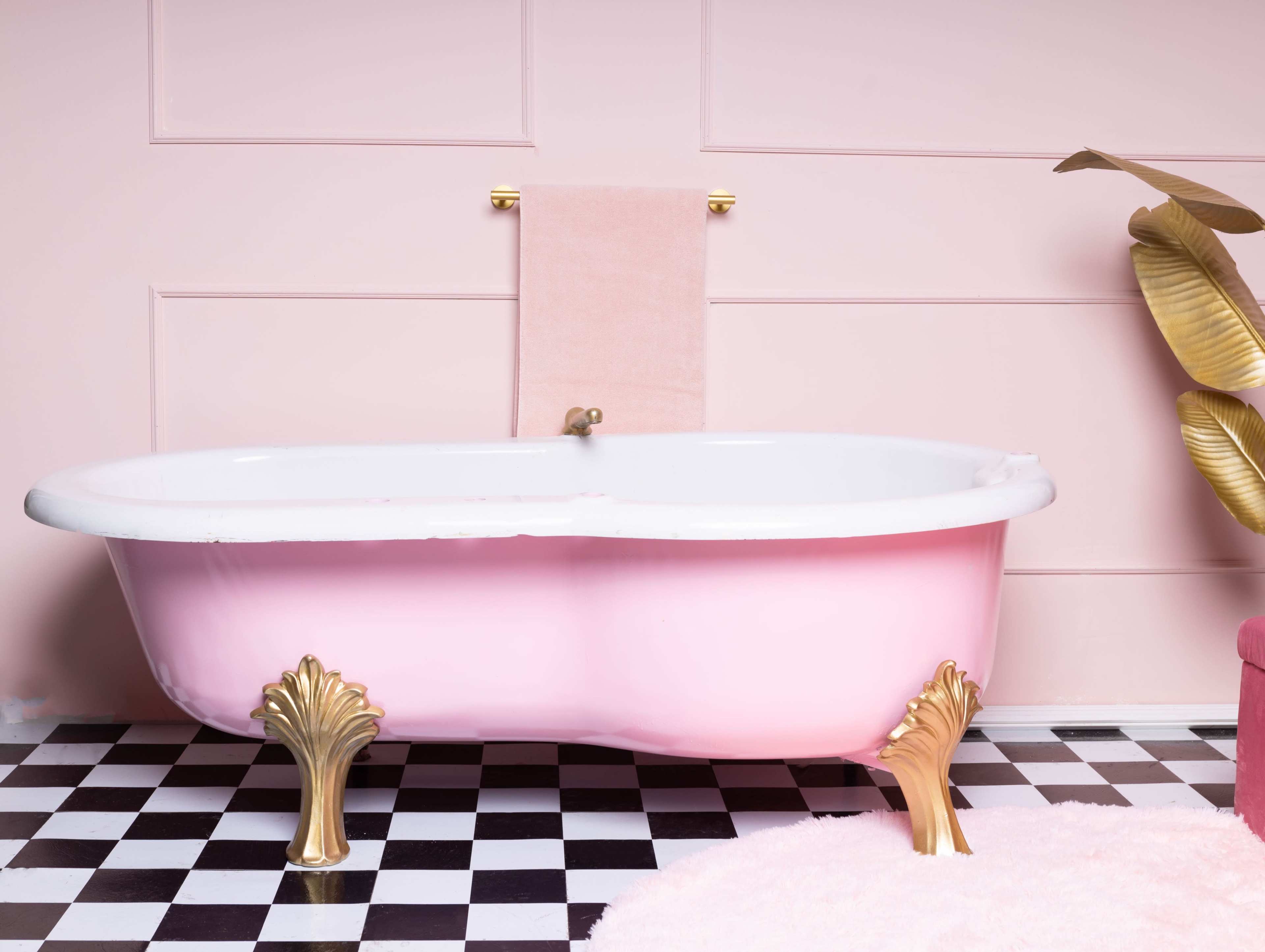 A pink freestanding bathtub with golden shell-shaped legs sits against a pink wall, accompanied by a towel rack and a plant in a modern setting with black and white checkered flooring.
