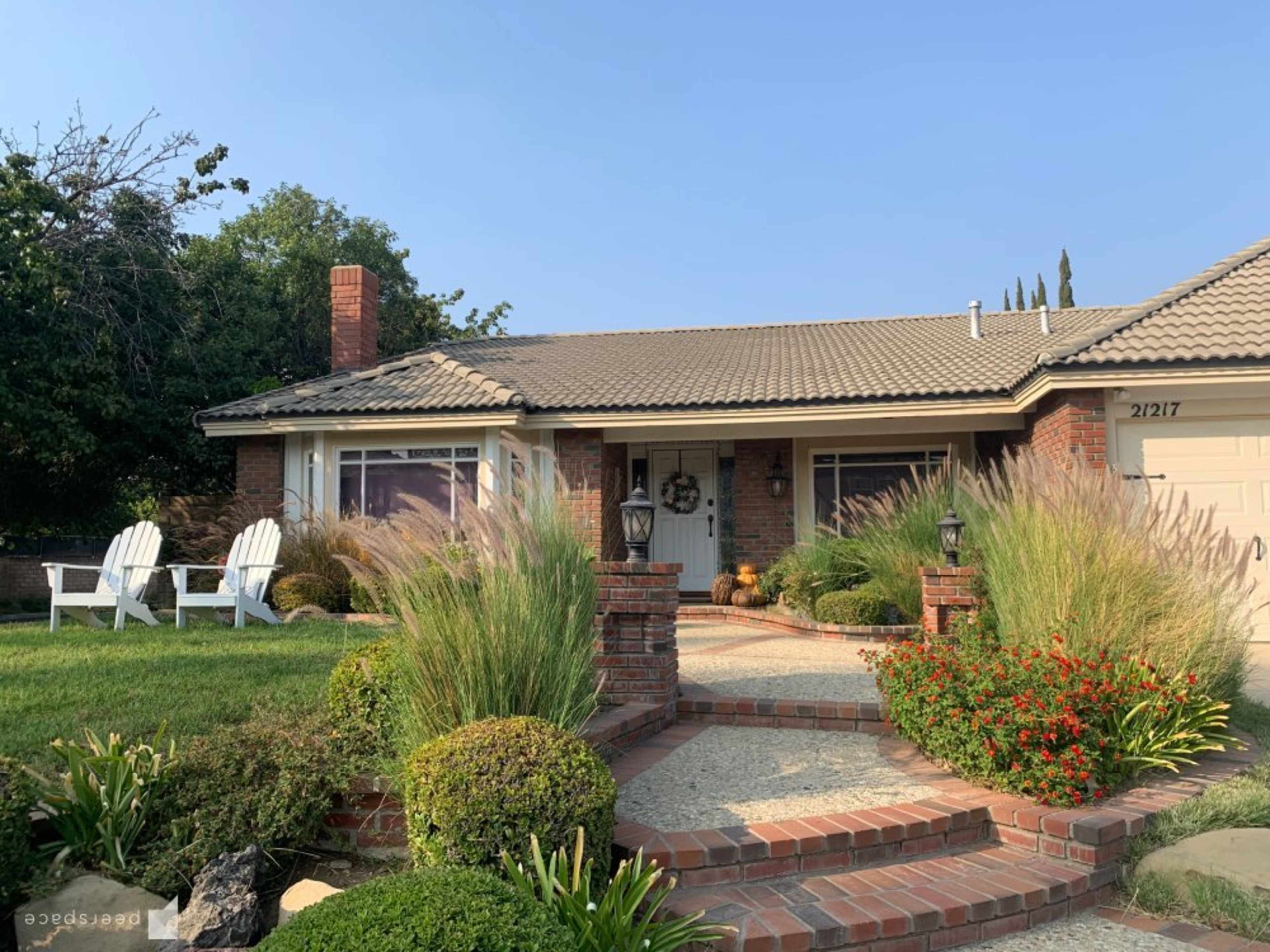 A single-story house with a brick facade, a tiled roof, and a landscaped front yard featuring shrubs and ornamental grasses.