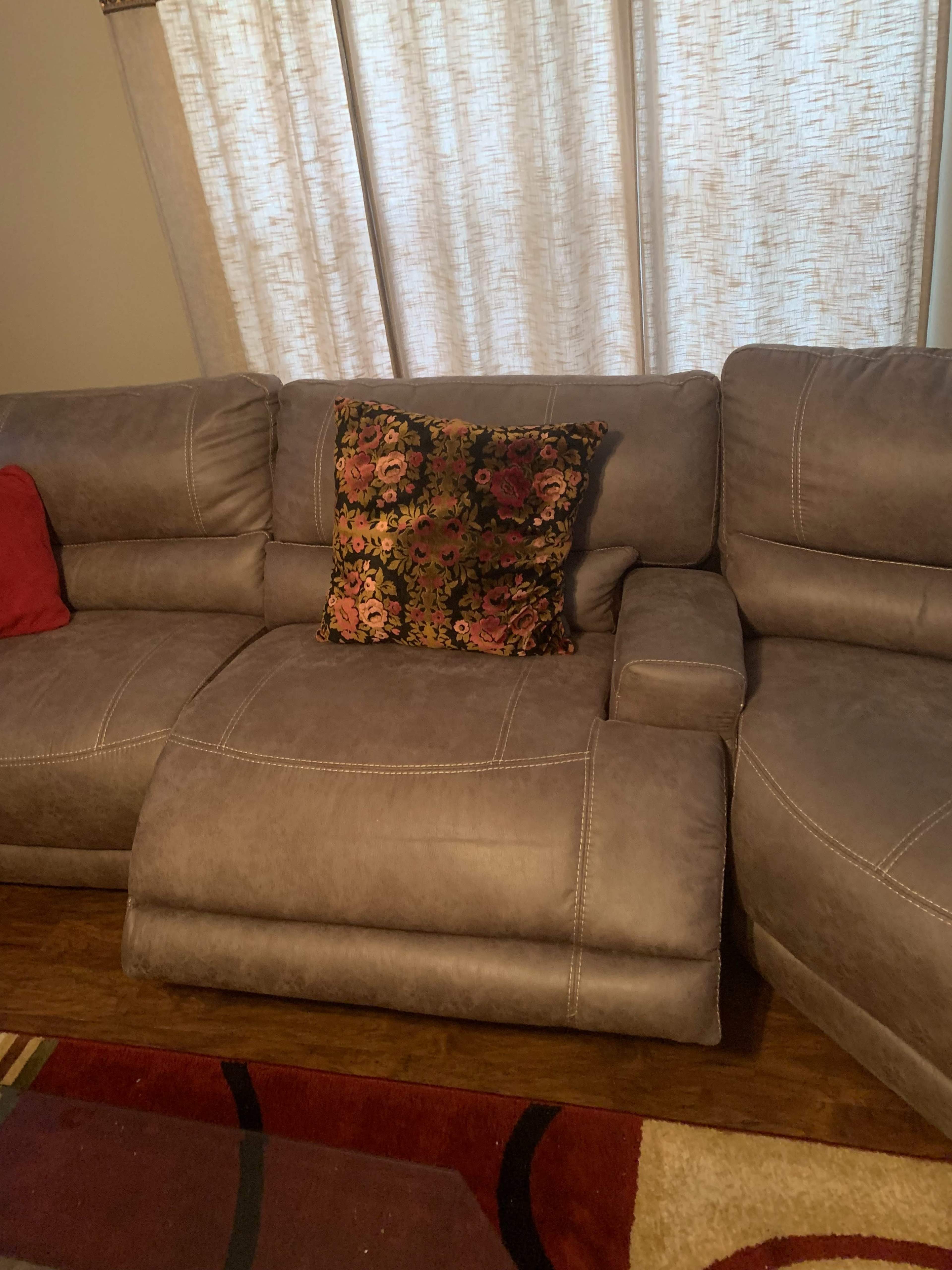 A sectional sofa with a floral pillow positioned on the corner seat, set against a backdrop of partially closed curtains.