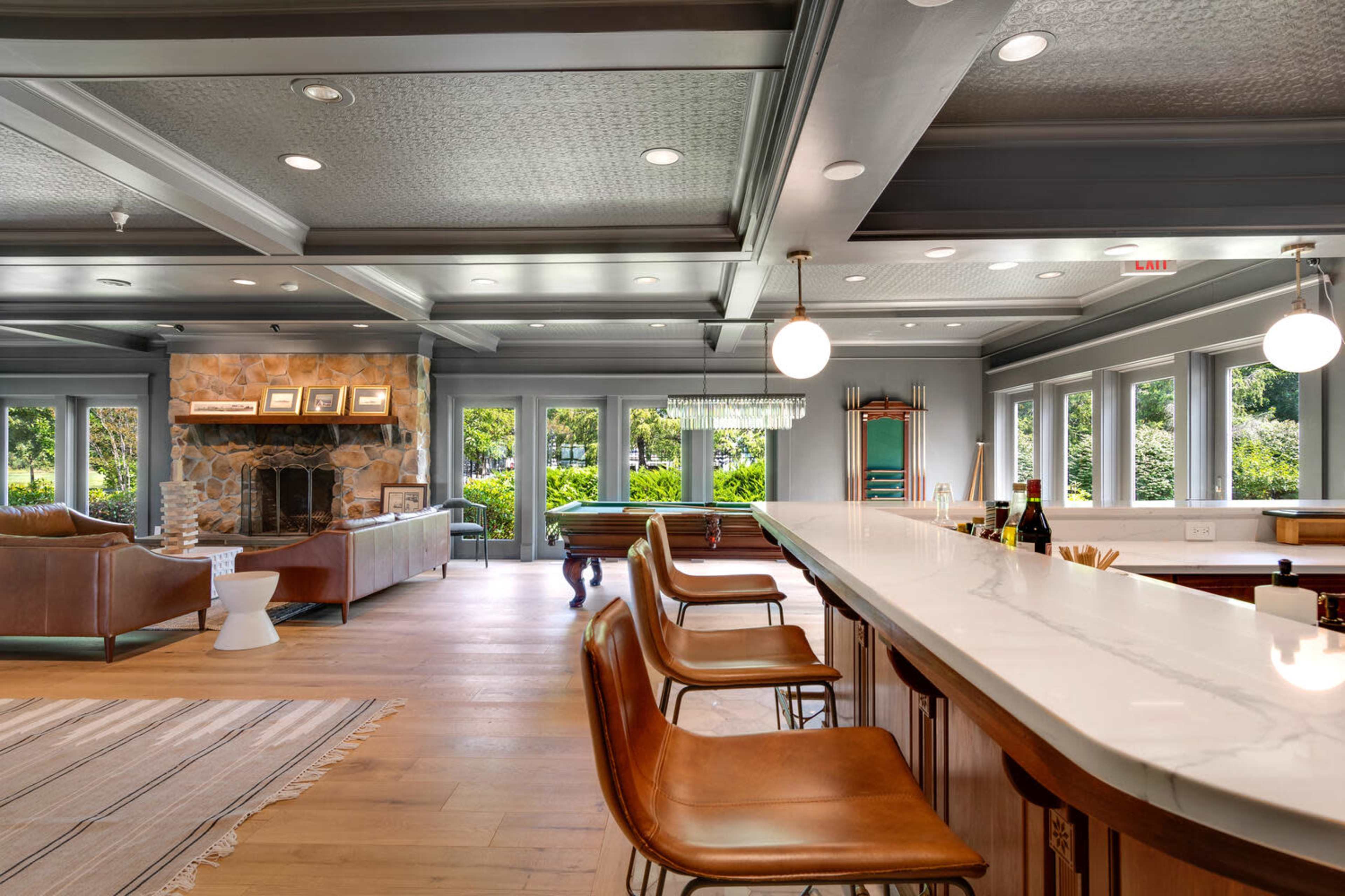 The image shows a spacious interior living area with a kitchen bar featuring four brown stools, a stone fireplace, and large windows overlooking greenery.