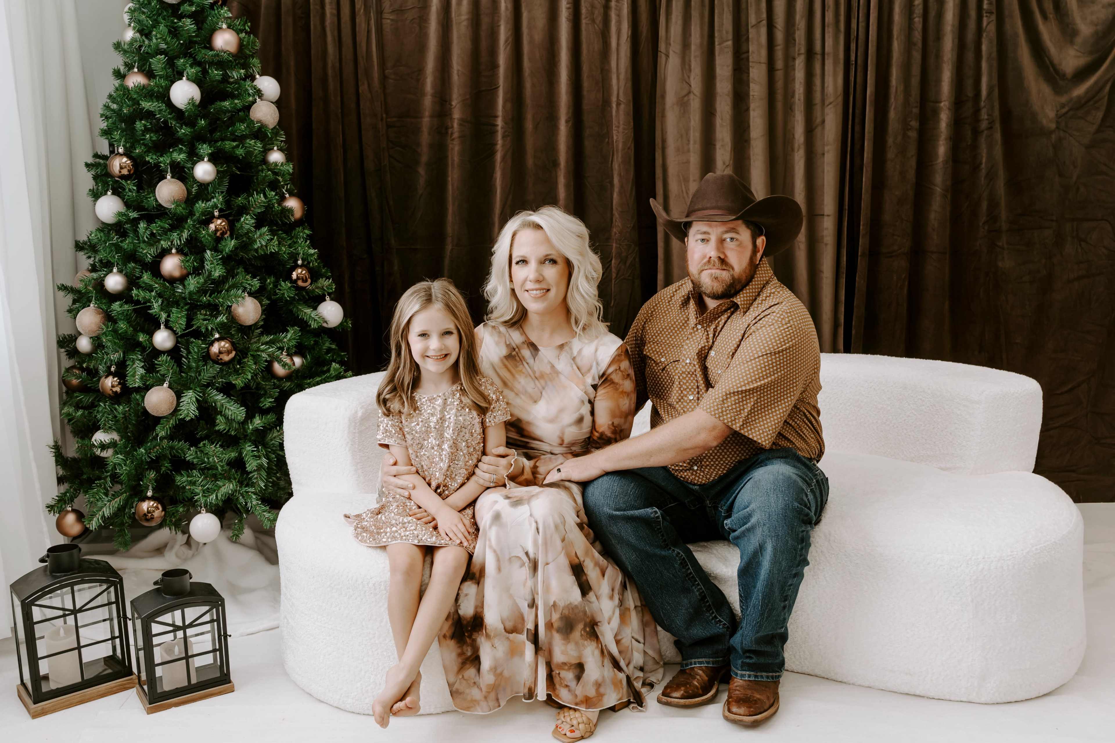 A family of three, including a woman, a man wearing a cowboy hat, and a young girl, sits together on a white sofa next to a decorated Christmas tree and lanterns.
