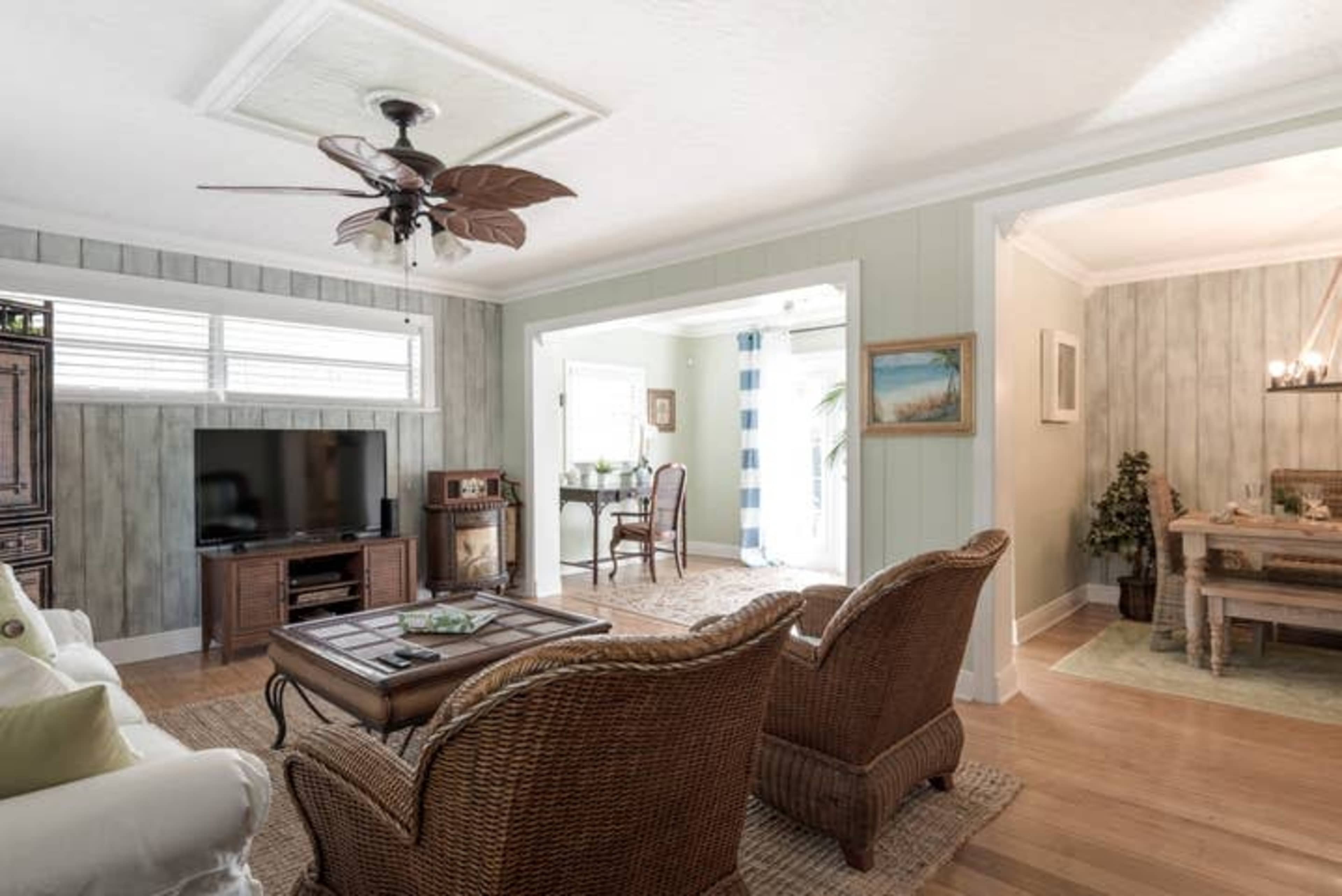 A well-lit living room with two wicker chairs facing a coffee table and a television, leading to an adjacent dining area with a small table and chairs.
