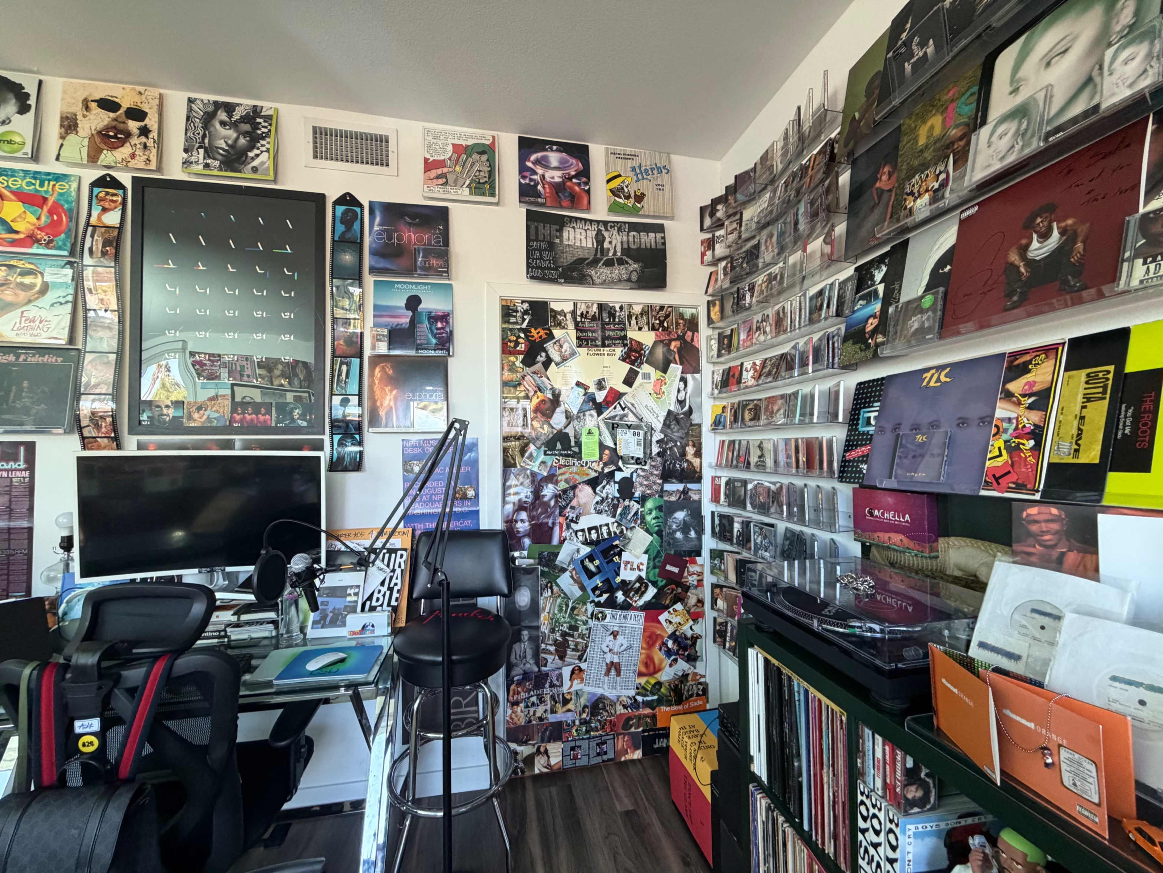 The image shows a music-themed room filled with posters, album covers, and vinyl records, featuring a desk set up for computer use and a barstool.
